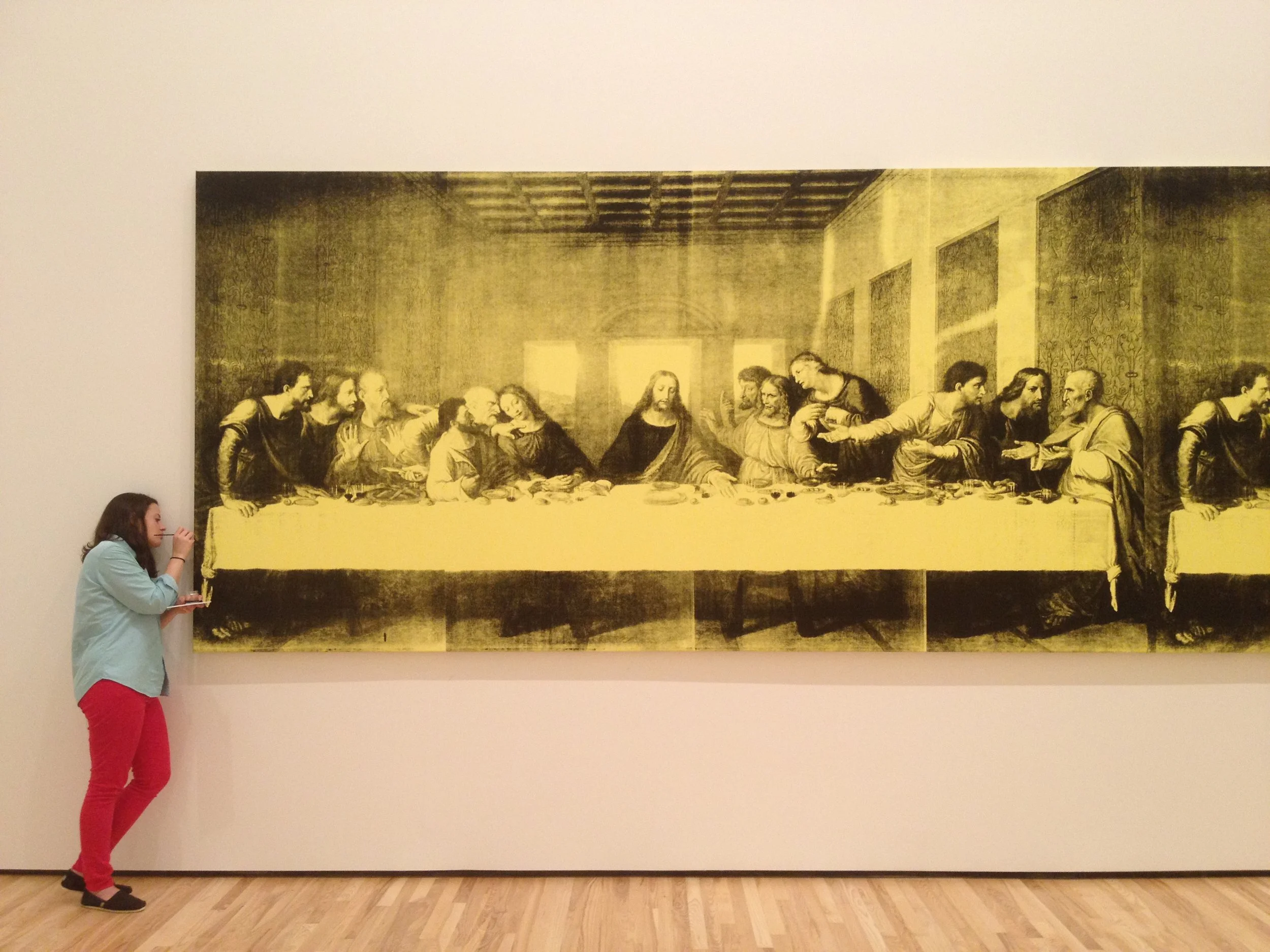 A woman standing to the left, looking at a large painting on a gallery wall that depicts the Last Supper.