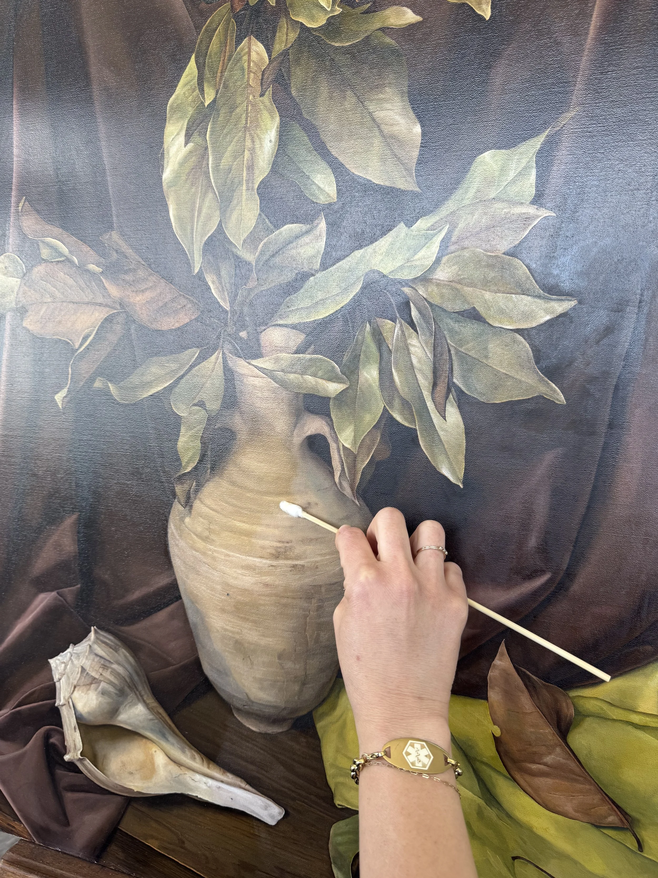 A person holding a long stick in front of a still life painting of a vase with green and brown leaves.