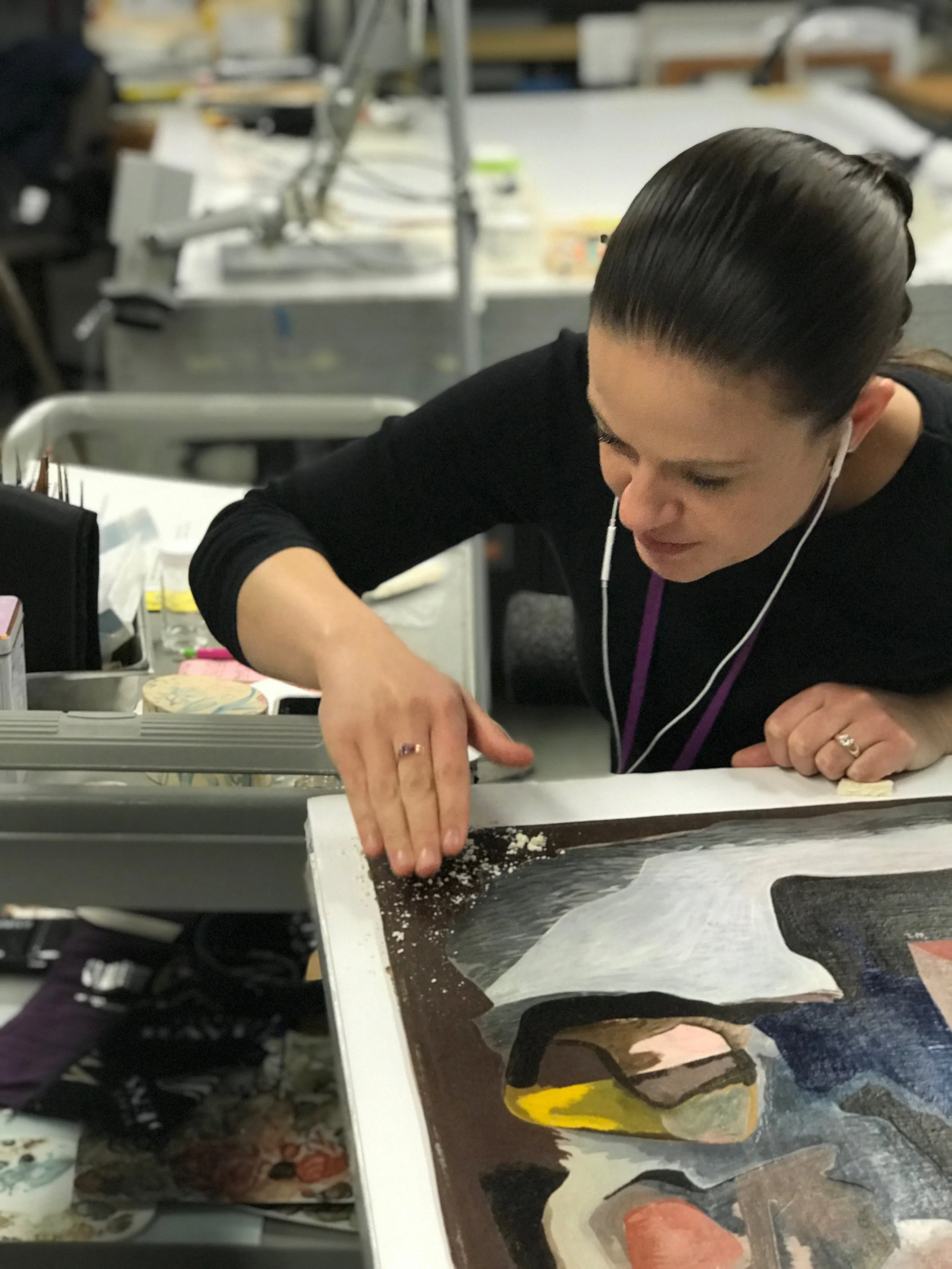 A woman with dark hair slicked back, wearing a black shirt and earbuds, is carefully working on a painting or print that depicts a figure in yellow, black, and white, on a studio worktable surrounded by art supplies and tools.