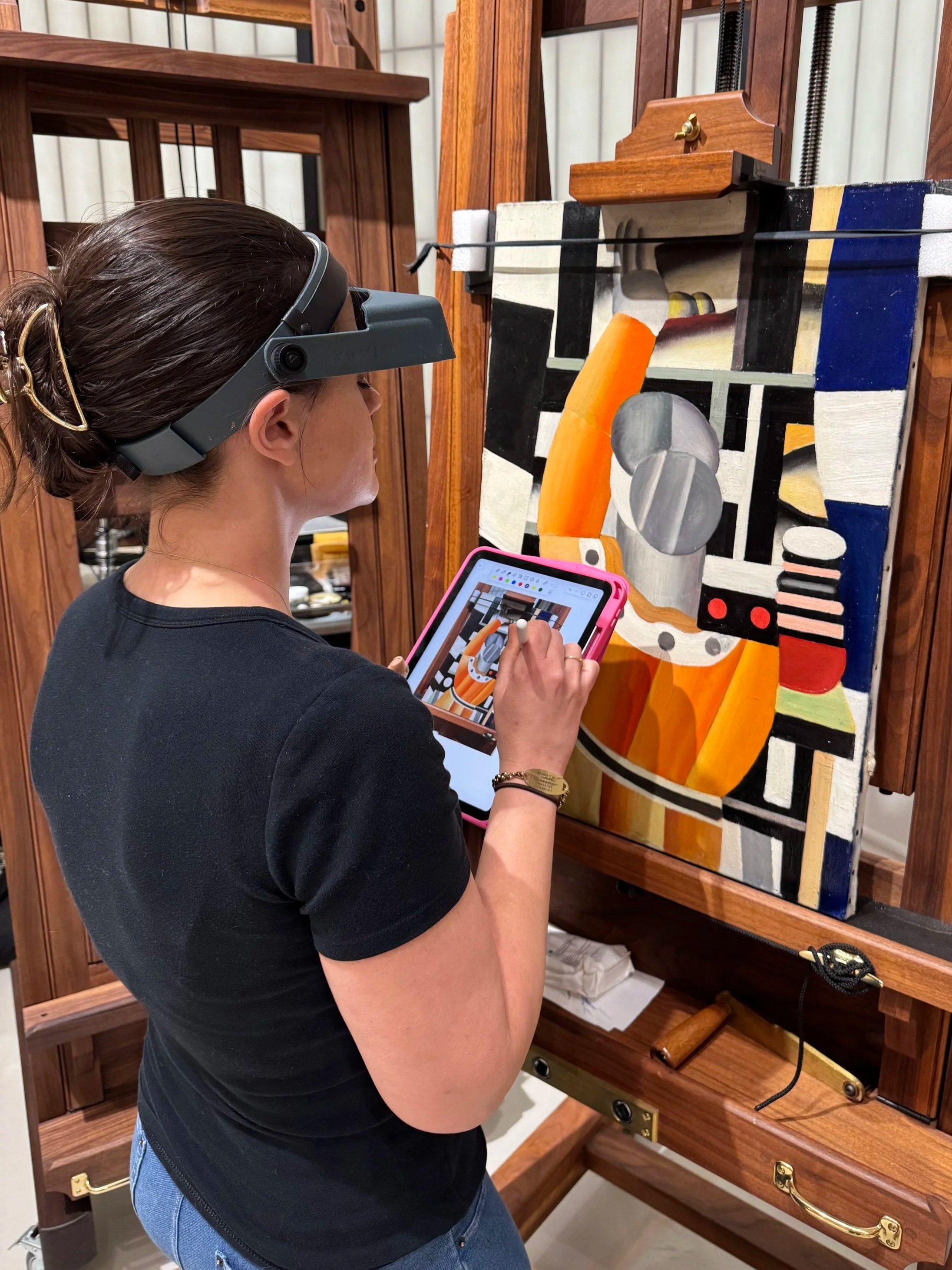 A woman with dark hair tied back, wearing glasses and a black shirt, is using a tablet while standing in front of a wooden antique mechanical music box or player piano.