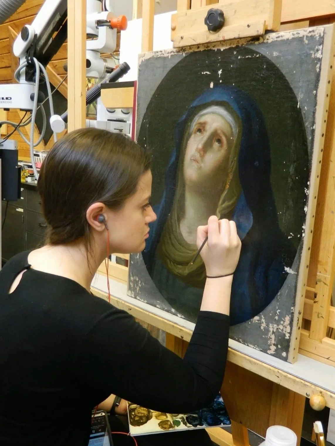A young woman with brown hair is restoring a classical religious oil painting of Virgin Mary, on an easel in an artist's studio, wearing a black shirt and earphones.