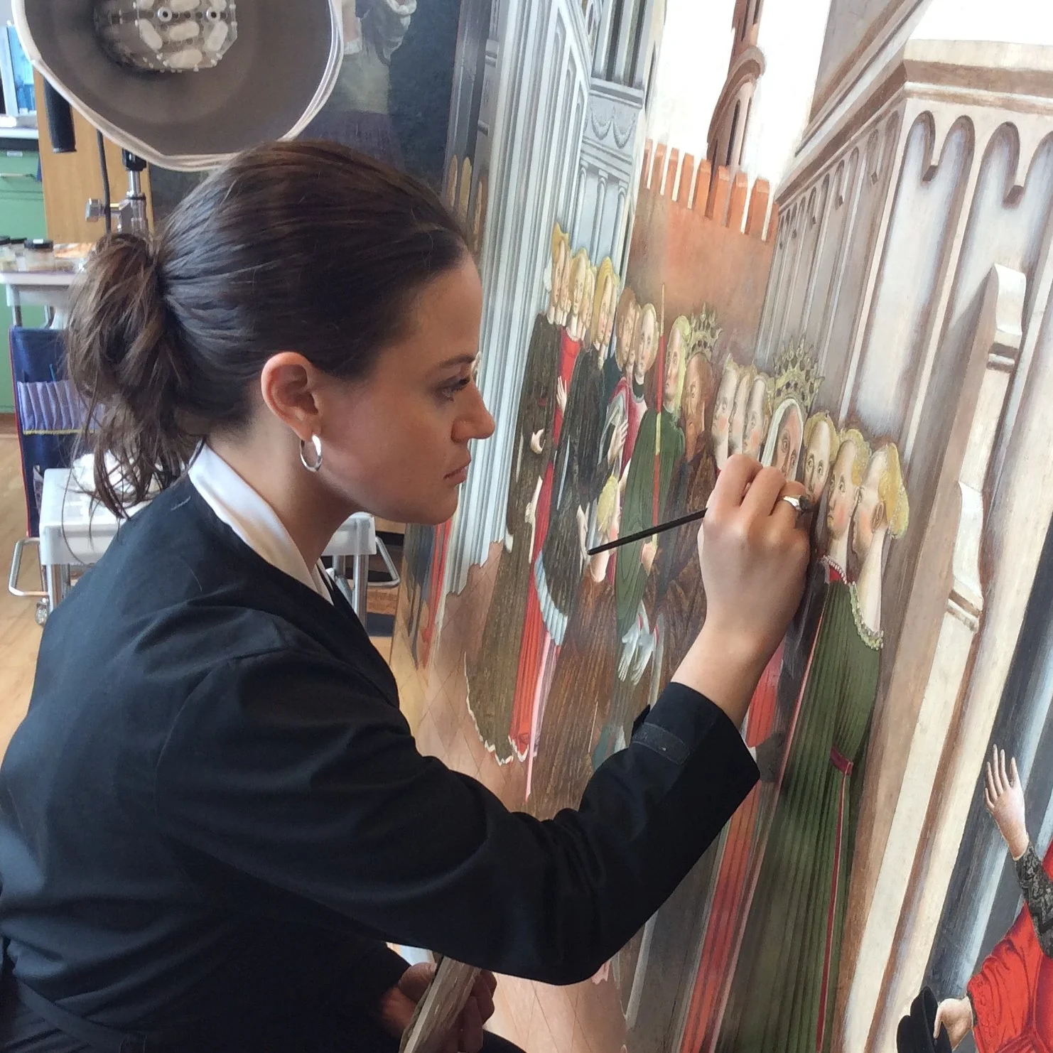 A woman with dark hair tied back wearing a black jacket and white shirt is treating an Italian panel painting.