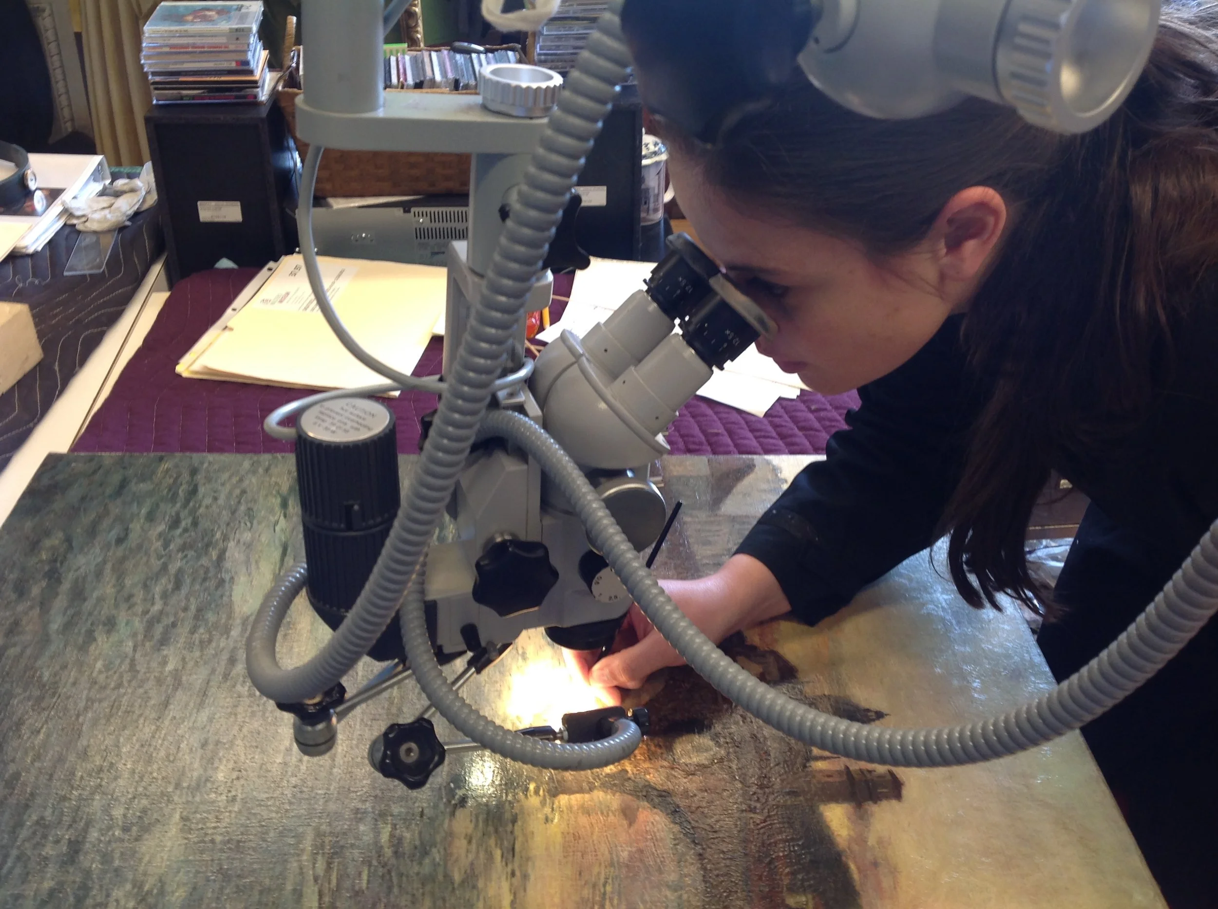 A person looking into a microscope at a laboratory or workshop setting, with equipment and papers on the table, surrounded by various supplies in the background.