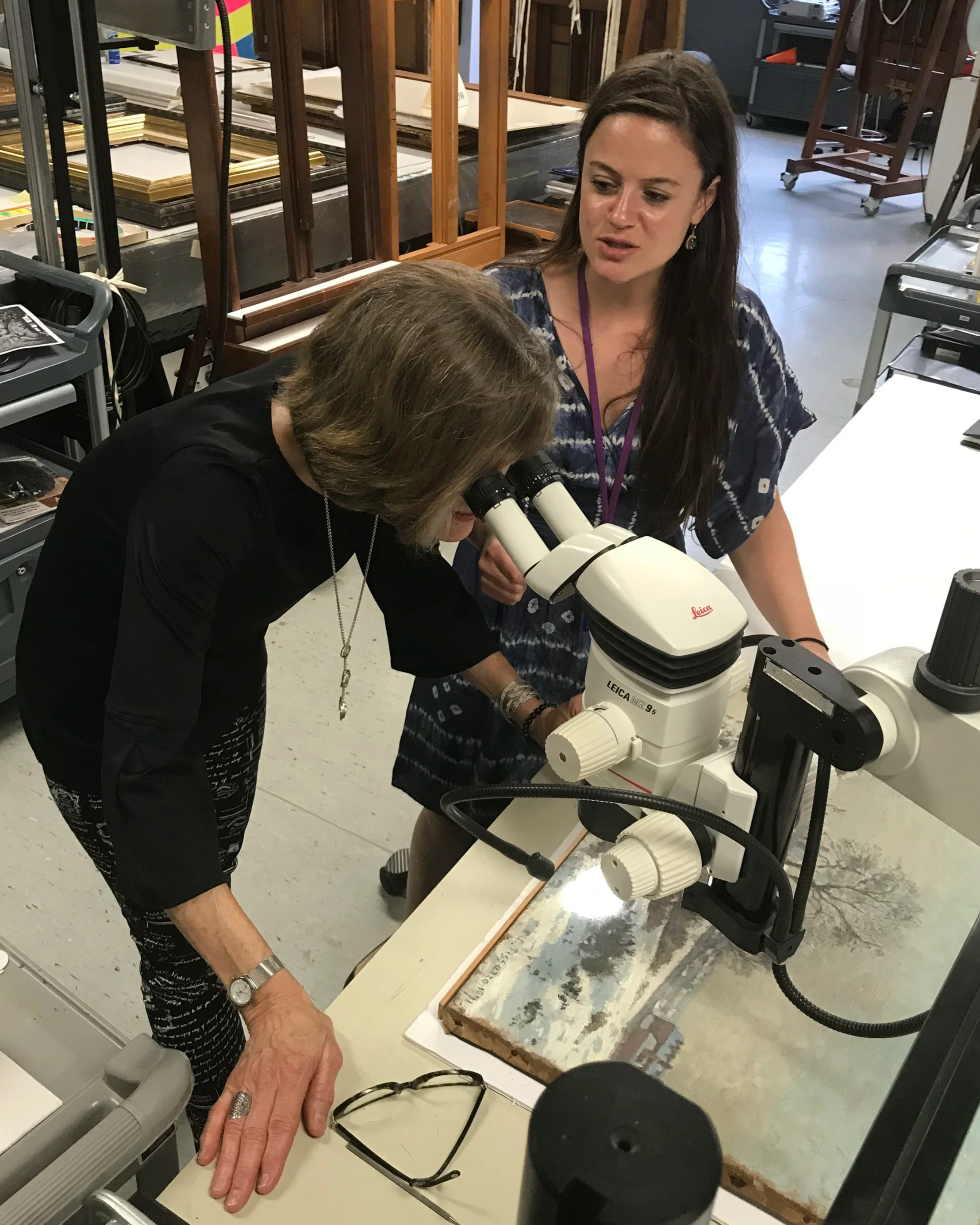 Two women are working with a microscope at a workspace with art supplies and frames in the background.