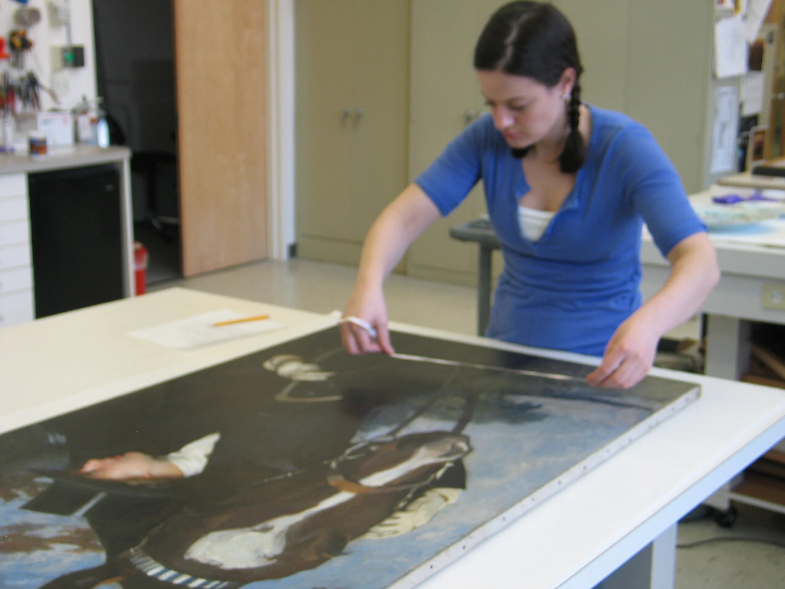 A woman with dark hair in braids working on an art project at a table in a room with cabinets and art supplies.