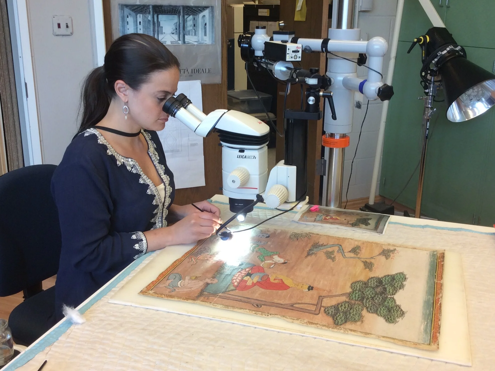 A woman with dark hair pulled back, wearing earrings and a dark jacket with a white shirt, looks into a microscope while working on a detailed, colorful book or artwork on a large table. The setting appears to be a laboratory or studio with scientific equipment and a poster on the wall.