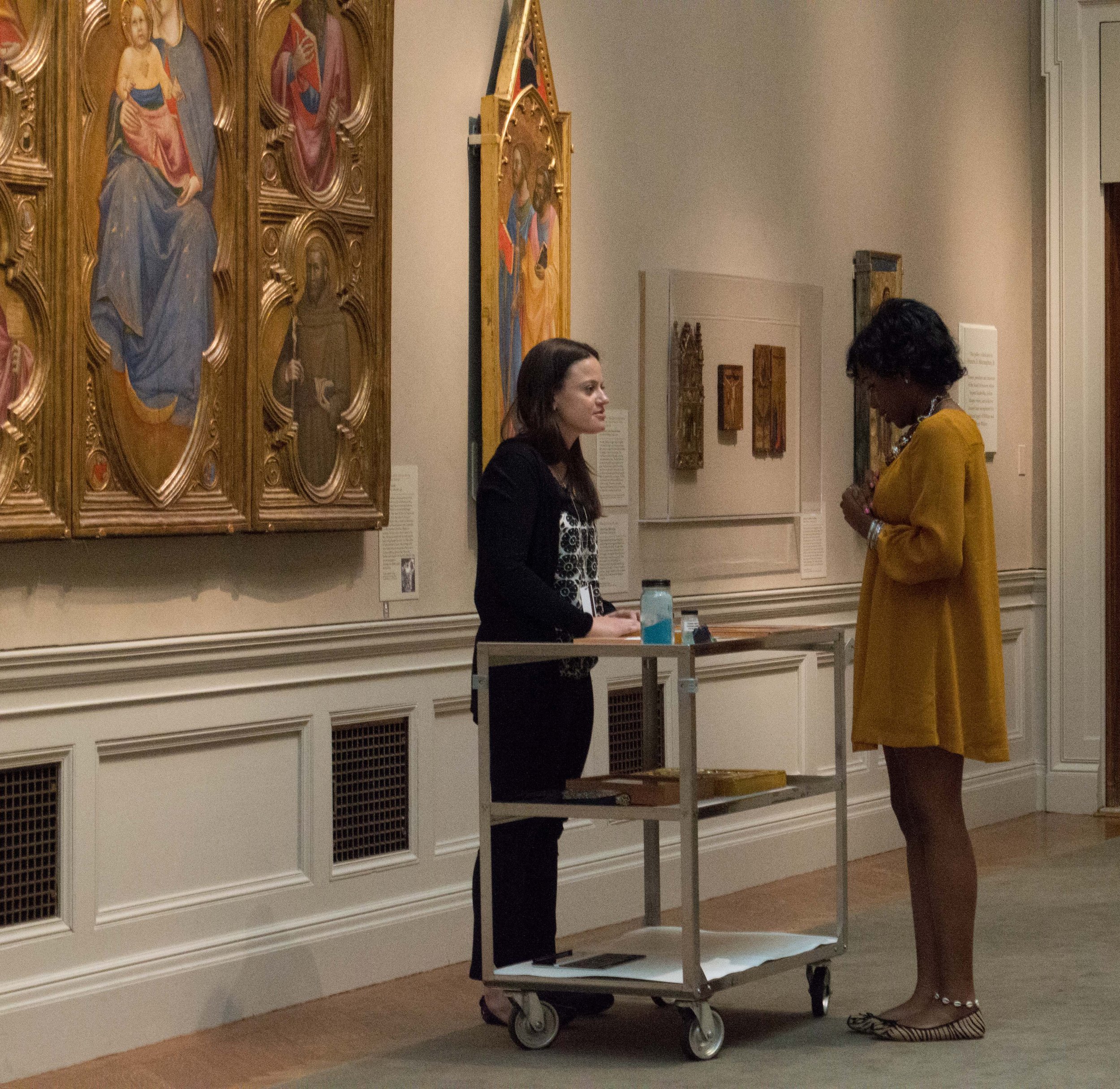 Two women are standing in an art gallery near religious paintings on the wall. One is dressed in black with long dark hair, the other in a mustard yellow dress with short curly hair. They are engaged in conversation with a table between them holding a water bottle and papers.