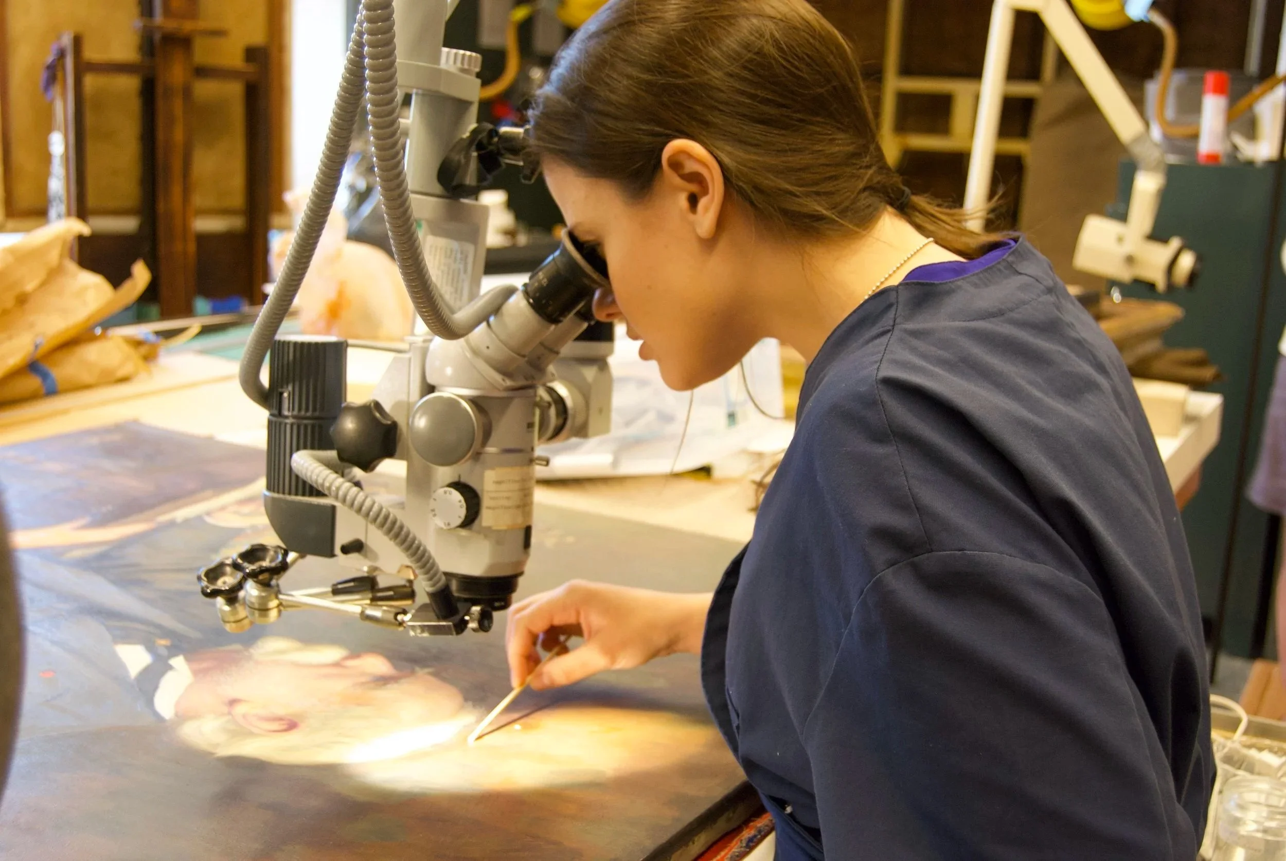 A woman using a microscope in a workspace with wooden shelves and materials nearby.