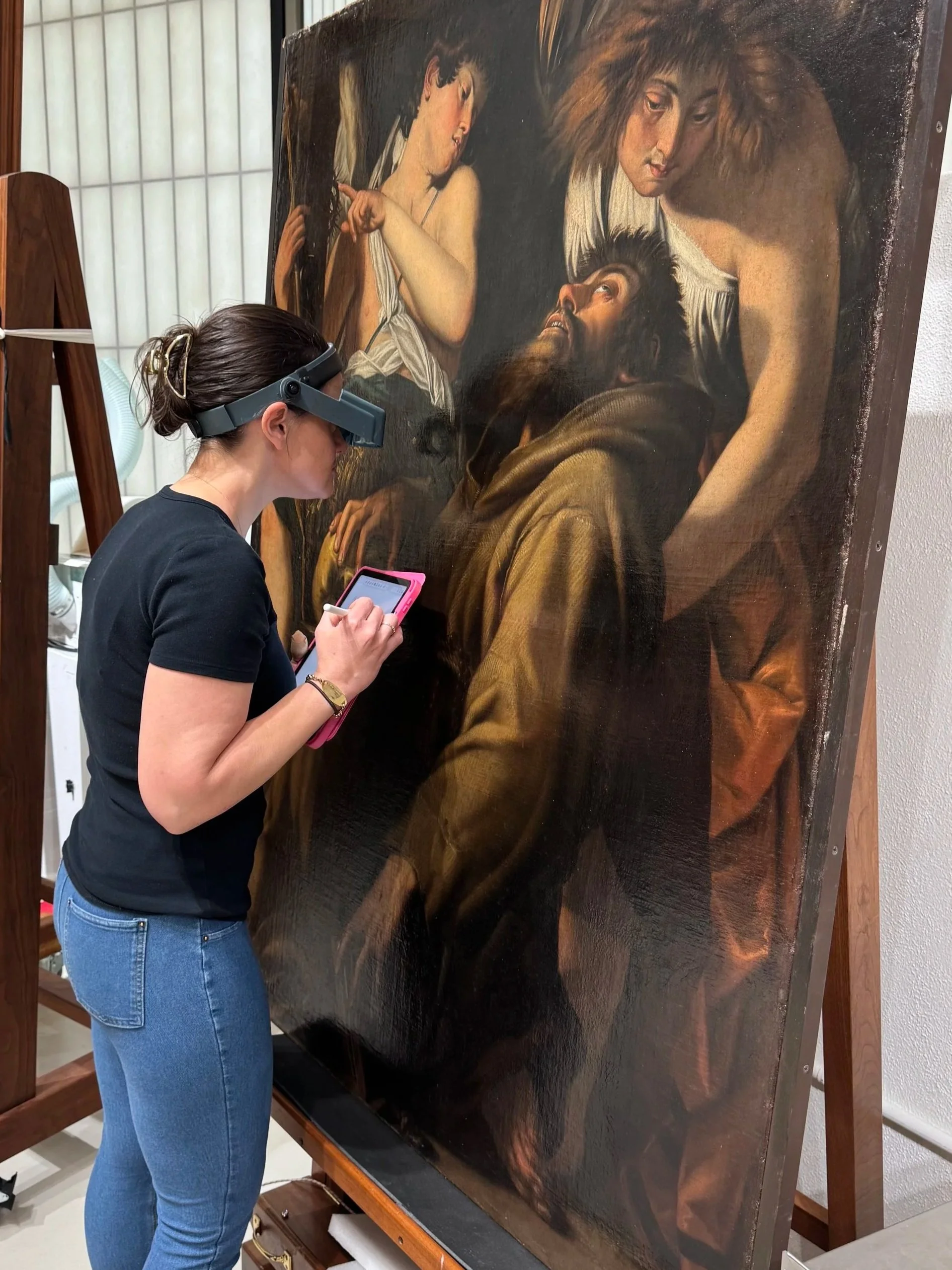 A woman wearing a head-mounted magnifier is examining a large, classical-style painting of Jesus Christ and two women, one of whom is pointing at Jesus.
