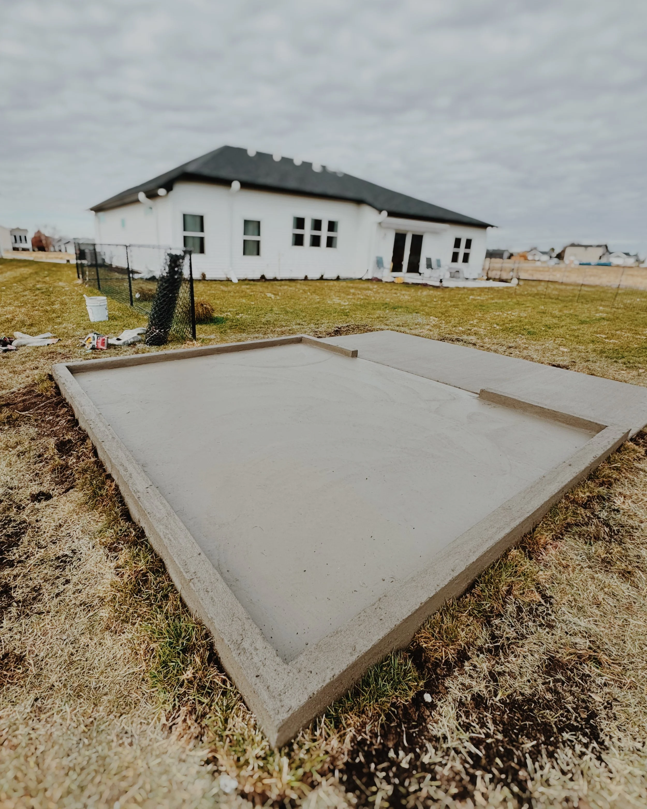 Newly poured cement slab in a backyard with a house and overcast sky in the background.