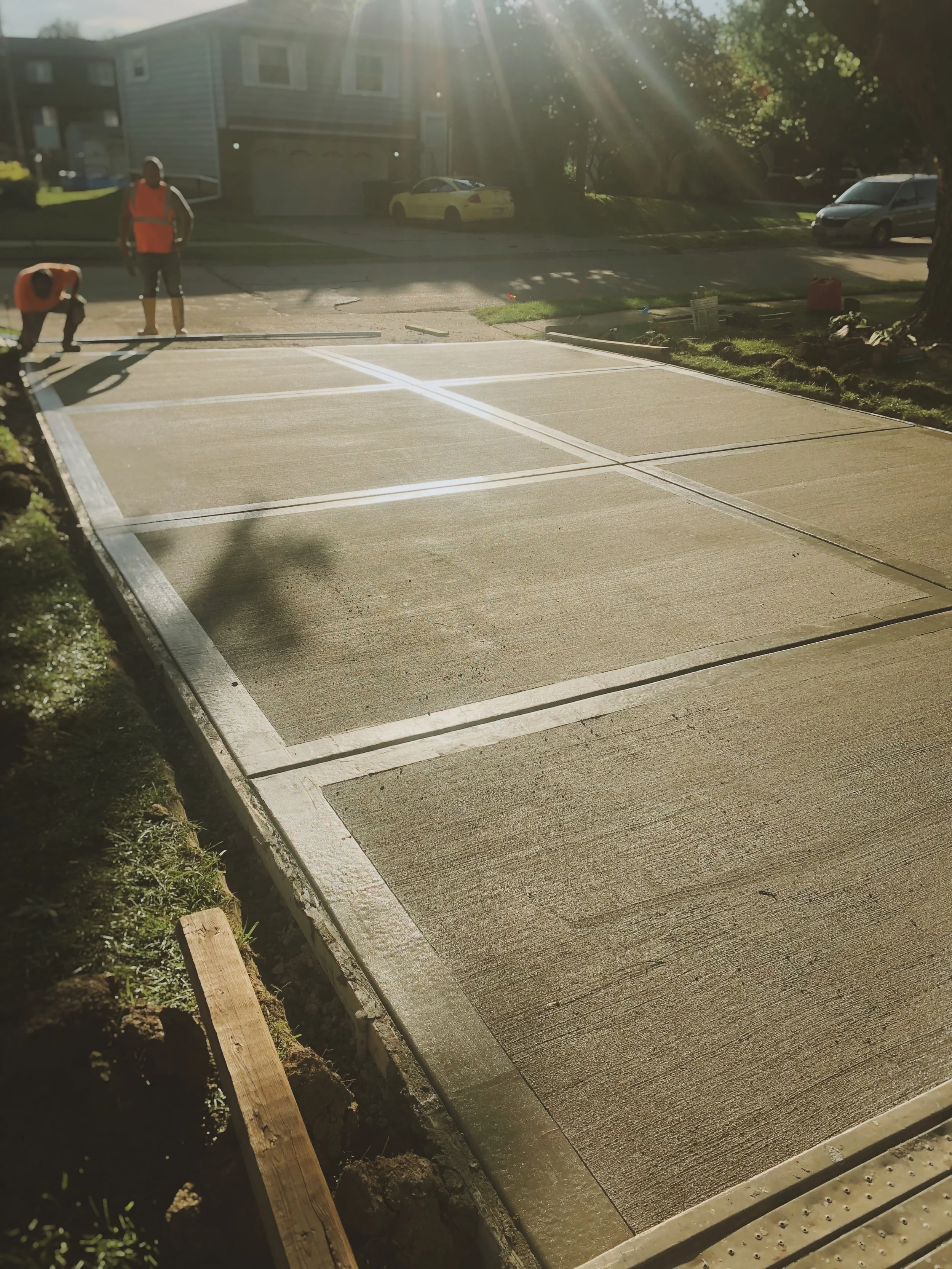Workers are laying down concrete for a driveway, with construction tools and materials nearby, in a residential neighborhood during sunny weather.