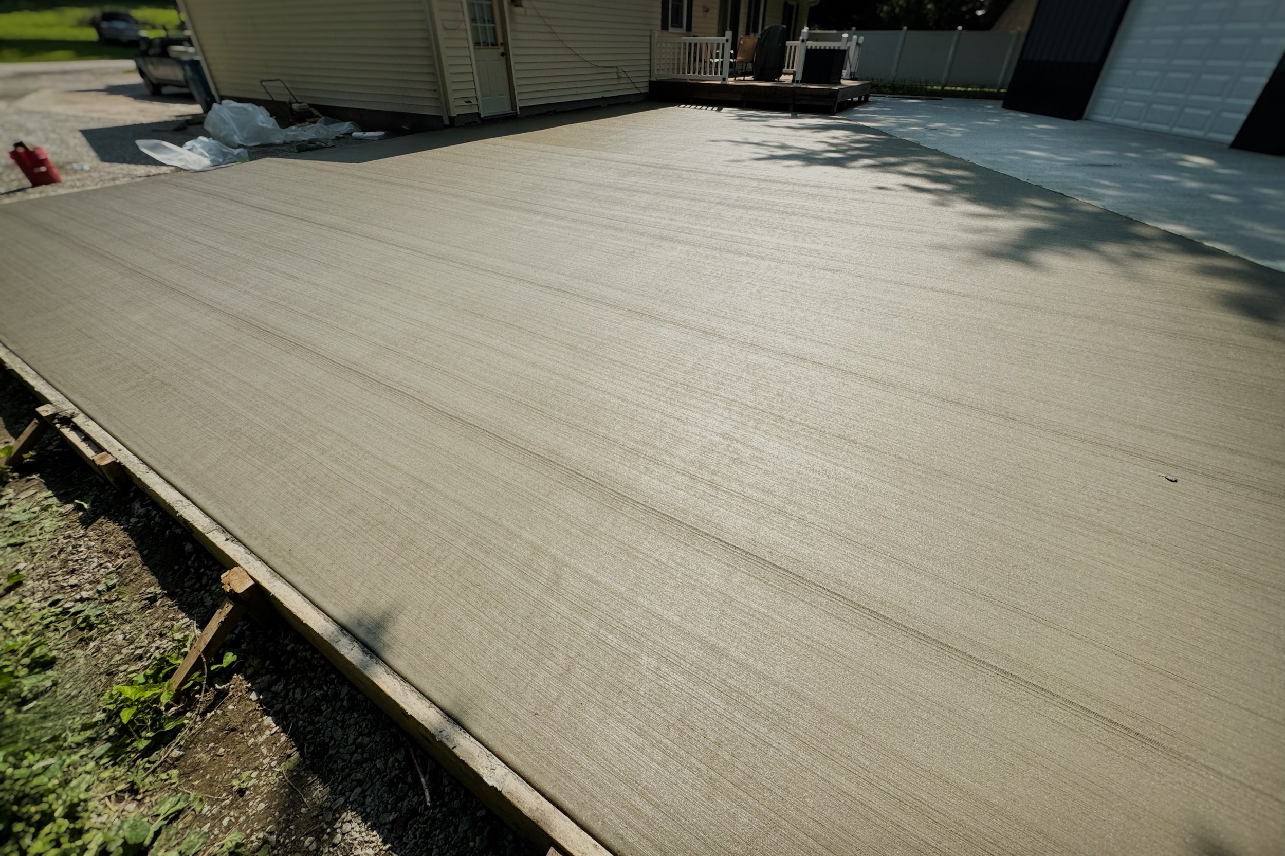 Freshly poured concrete slab in a backyard, with shadows of trees and a house in the background, surrounded by construction materials and tools.