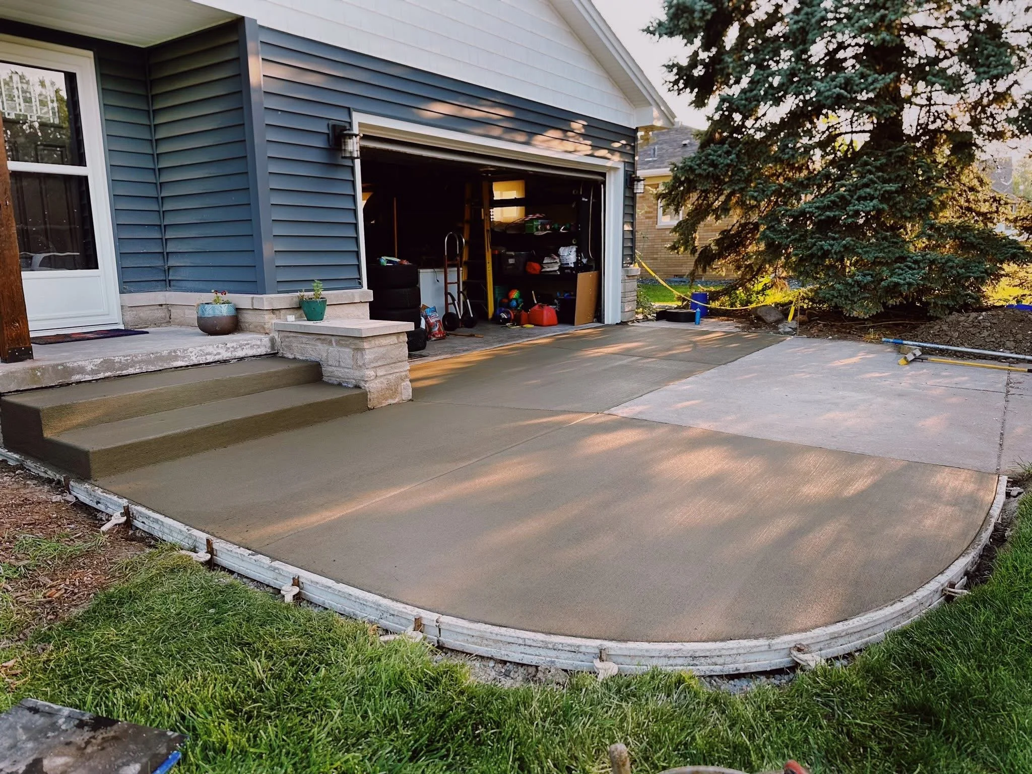 A newly poured concrete driveway in front of a house with siding, with a large tree on the right side and construction supplies nearby.