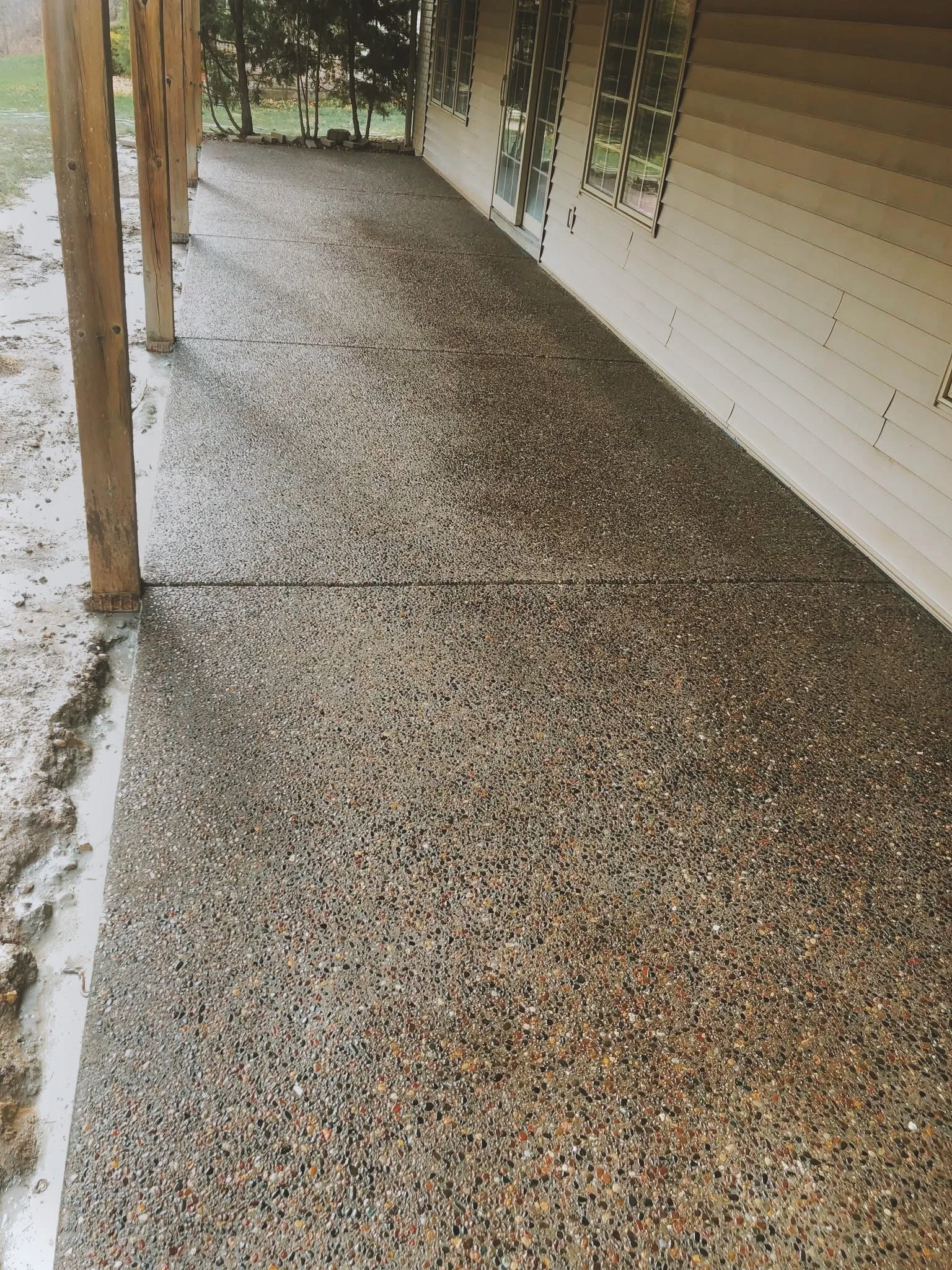 Concrete porch with pebble finish, attached to a house with beige siding and several windows, supported by wooden posts on the left, with some snow and dirt at the edge.
