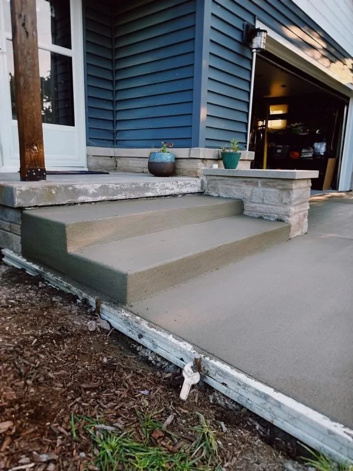 Newly poured concrete porch with two steps leading up to it, adjacent to a house with blue siding and a garage door open in the background, potted plants on the porch, and dirt at the base of the stairs.