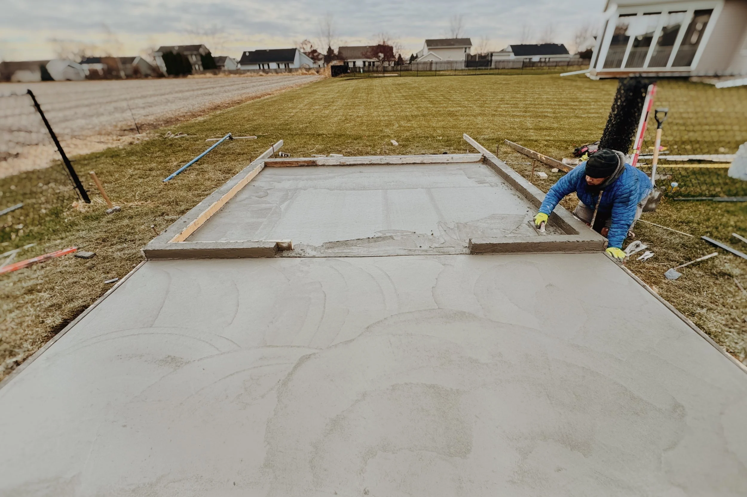 A person working on a concrete slab in a backyard, surrounded by tools and construction materials, with a grassy yard, houses in the background, and a partly cloudy sky.
