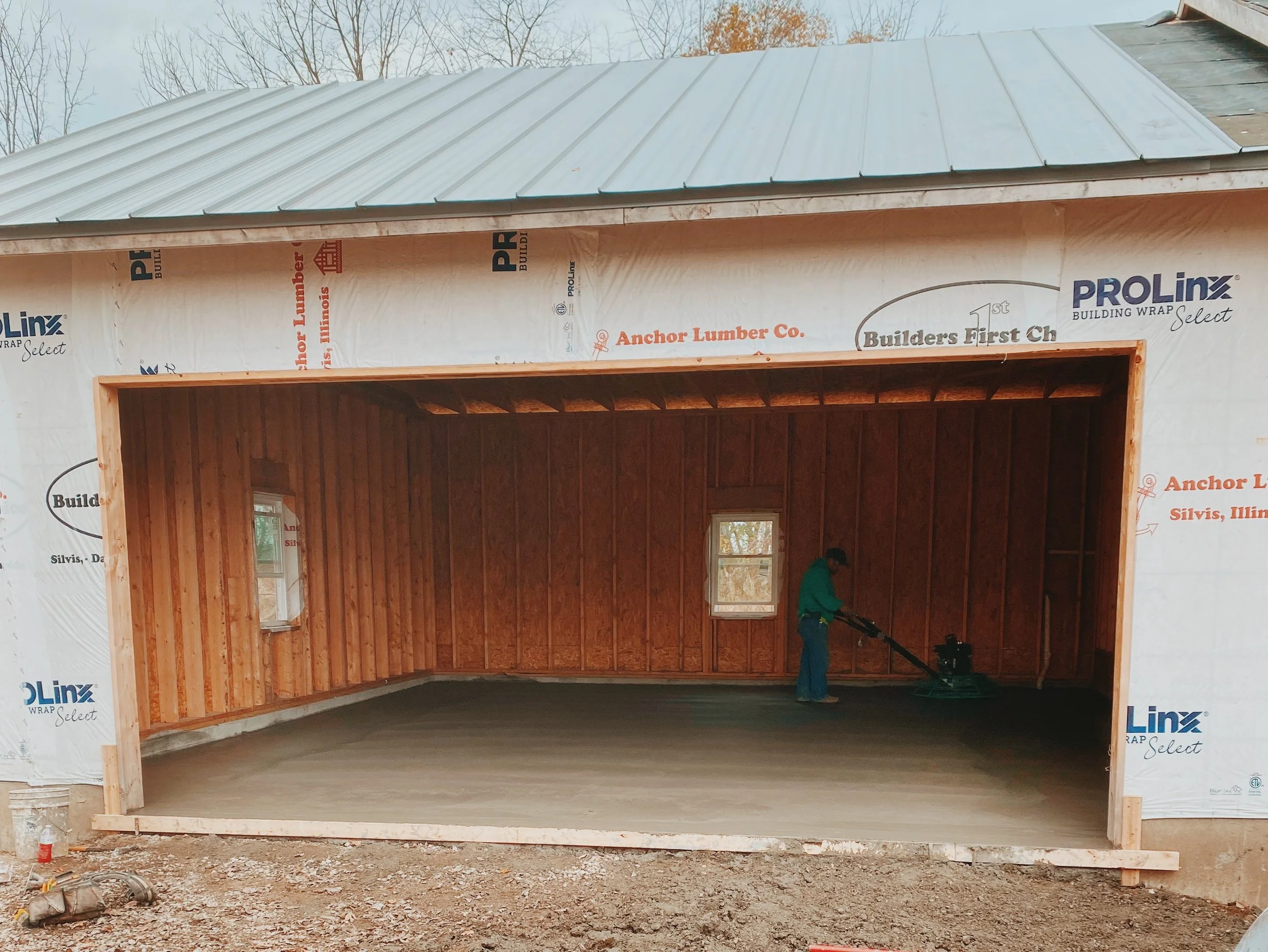 Construction site of a house with the walls covered in white builder's wrap and a metal roof installed, visible wooden framing, and a worker standing inside.