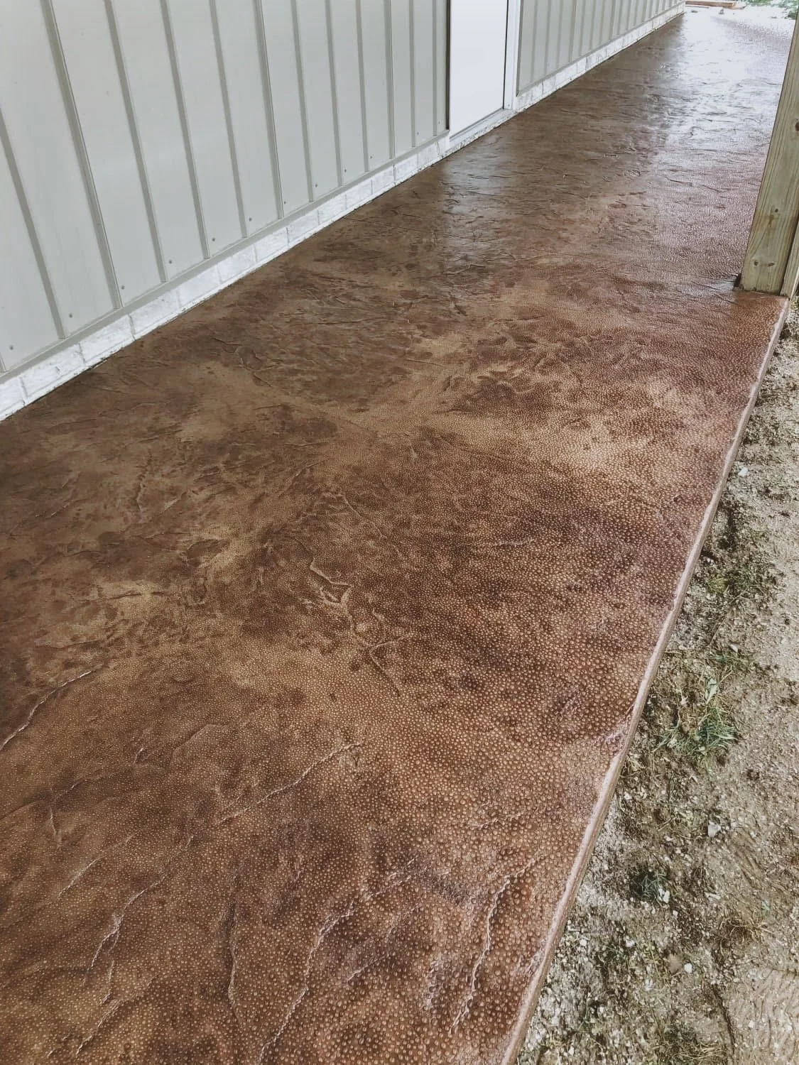 A brown concrete porch with a textured surface next to a gray metal building and a wooden support post.