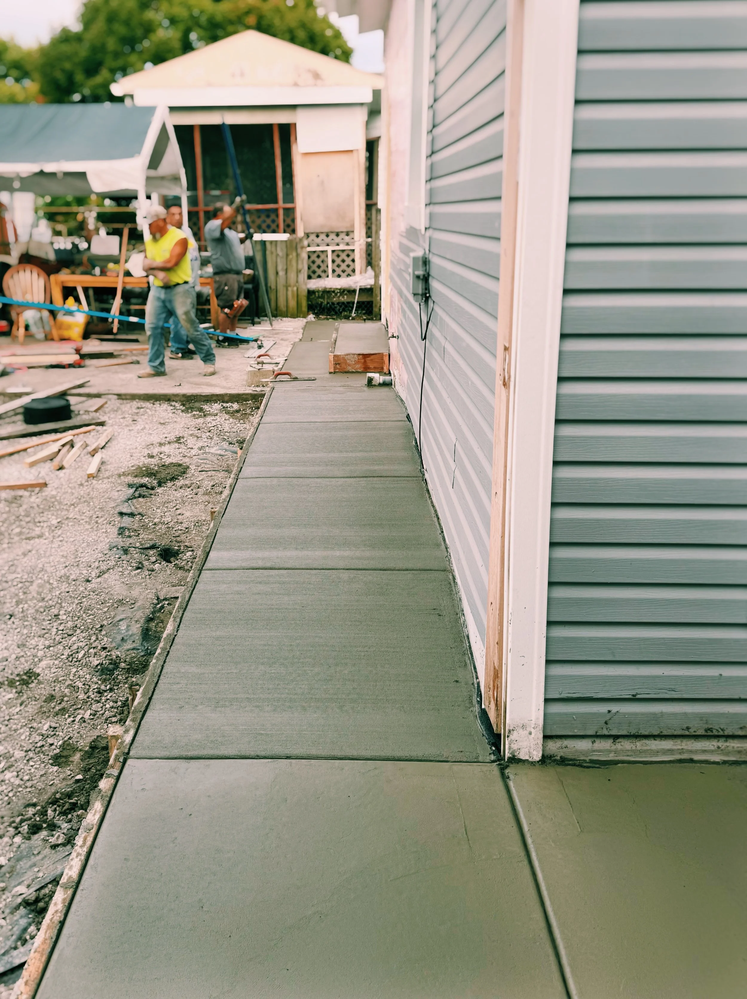 Workers pouring and finishing concrete sidewalk next to a house with blue siding, construction tools and materials scattered around.