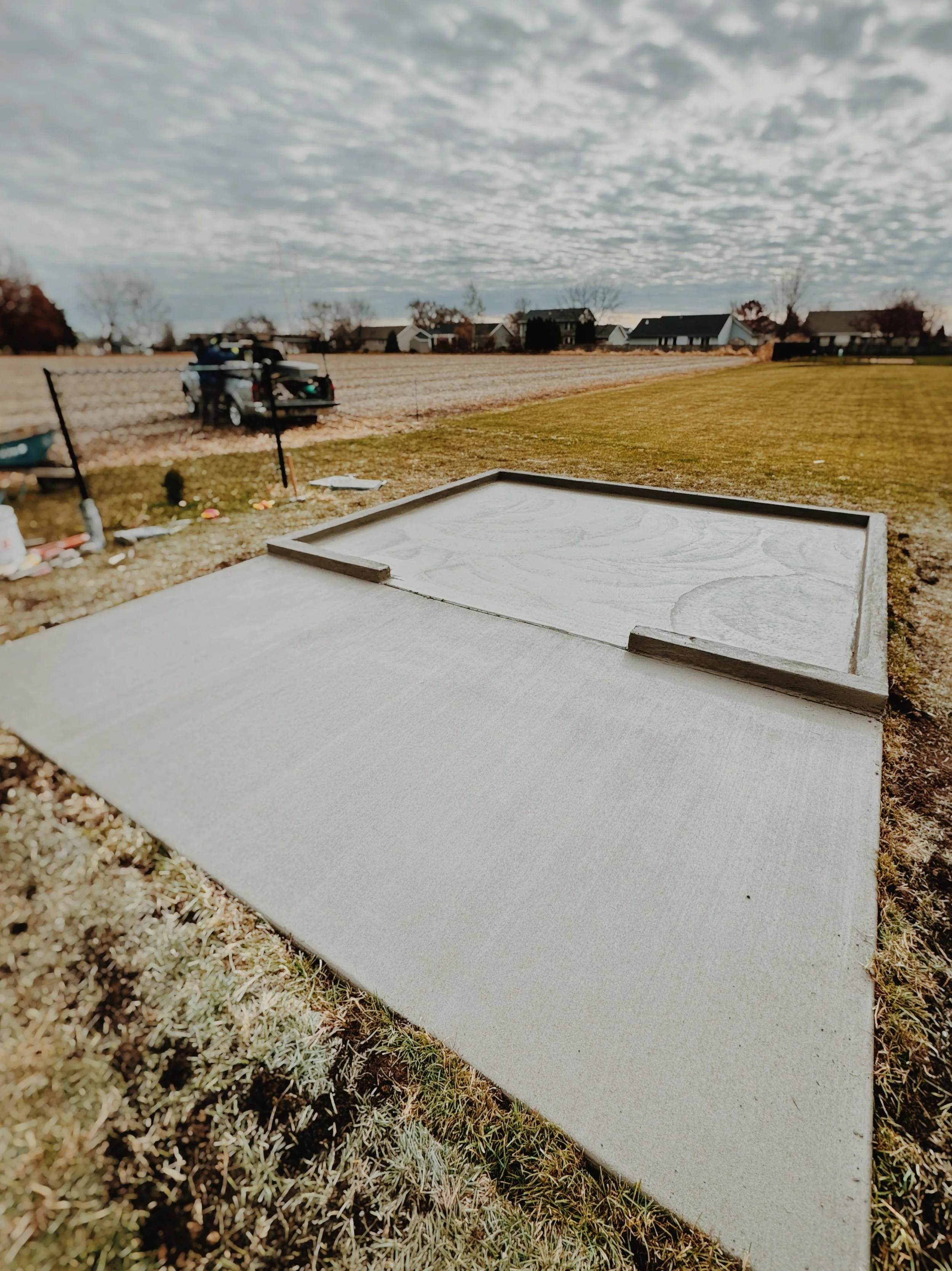 A wooden tray with a raised edge on a flat surface in a lawn area, with a background of a field, houses, and cloudy sky.