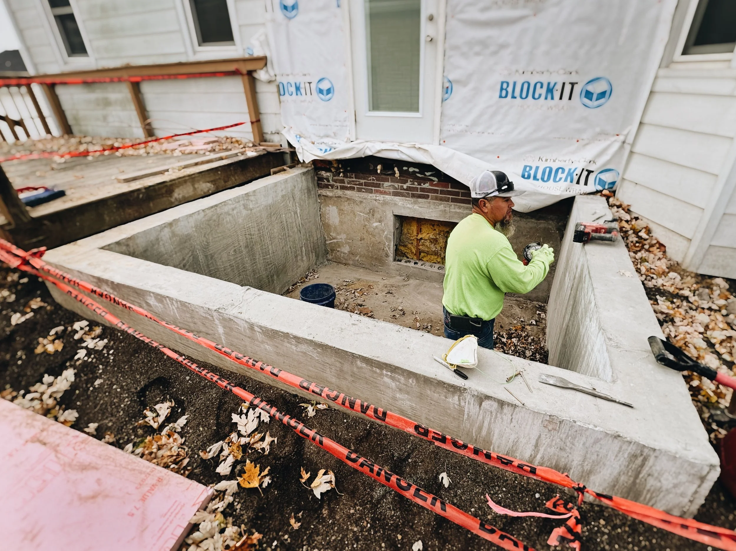 Construction worker in a neon yellow shirt working on a house foundation with damaged siding, surrounded by caution tape, leaves, and construction materials.