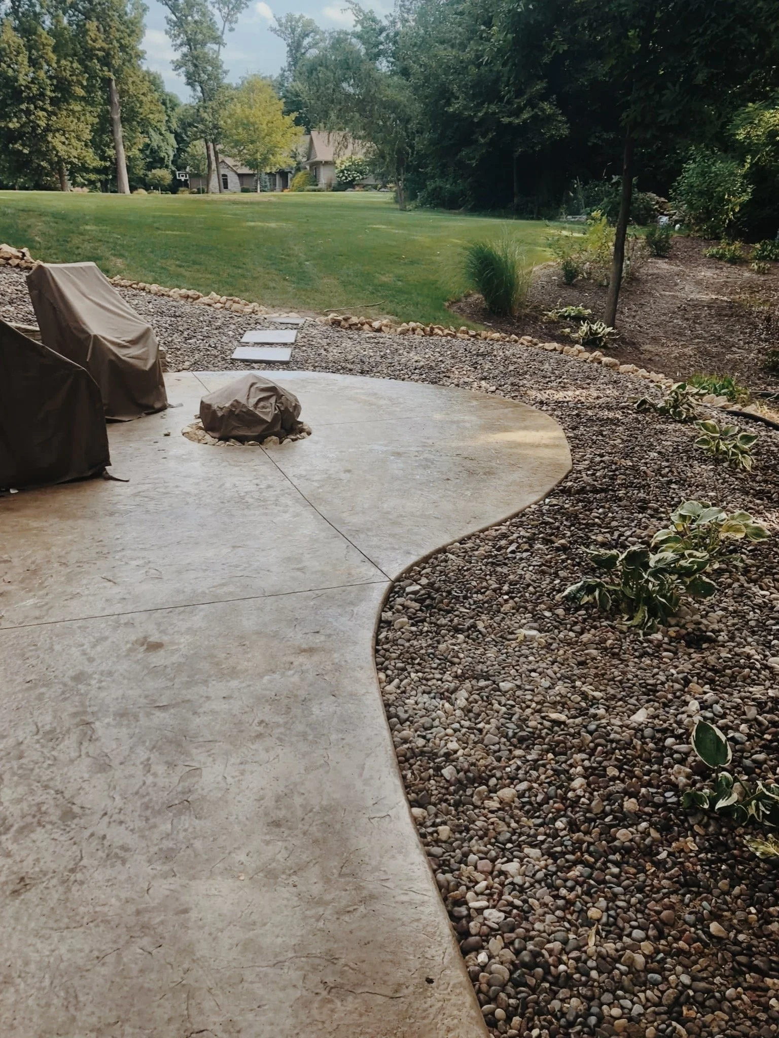 A backyard with a concrete patio, covered outdoor furniture, and a curved gravel border with plants. A lush green lawn extends into the background with trees and a house with a basketball hoop visible in the distance.