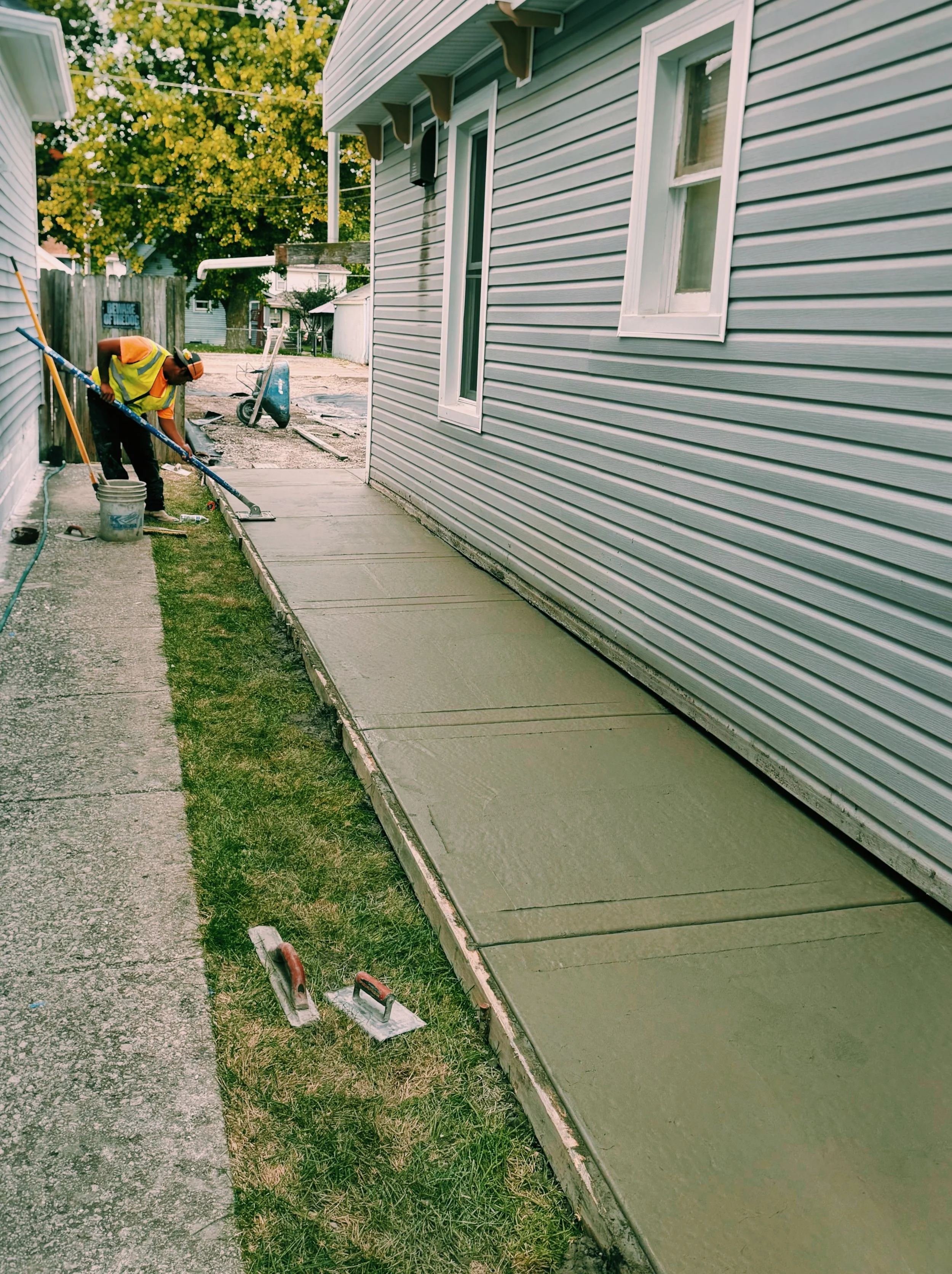 Worker applying fresh concrete to a sidewalk walkway next to a house with gray siding, using a long-handled tool, with construction tools nearby.