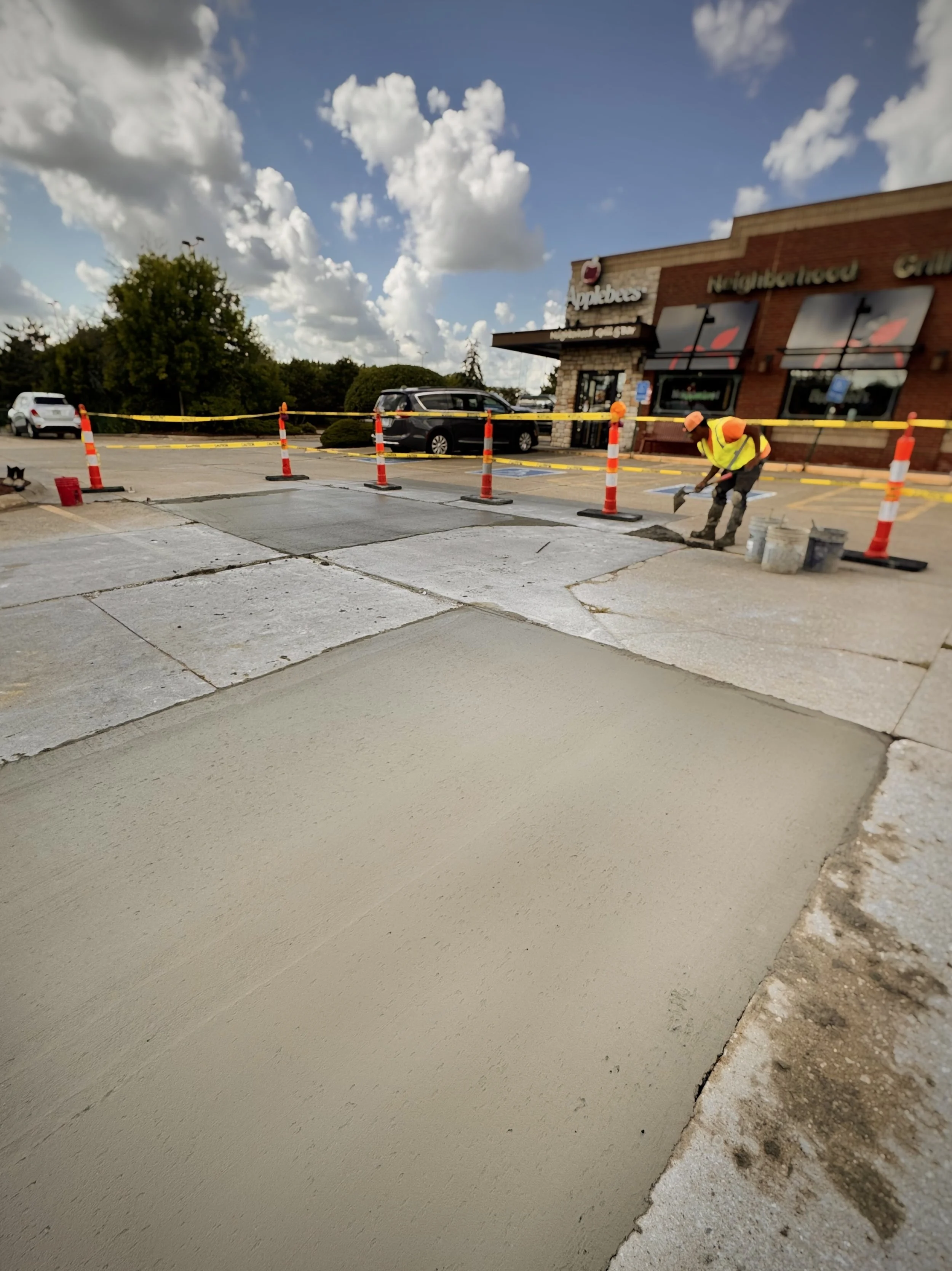 Worker in a high-visibility vest smoothing wet concrete in a parking lot outside a restaurant.