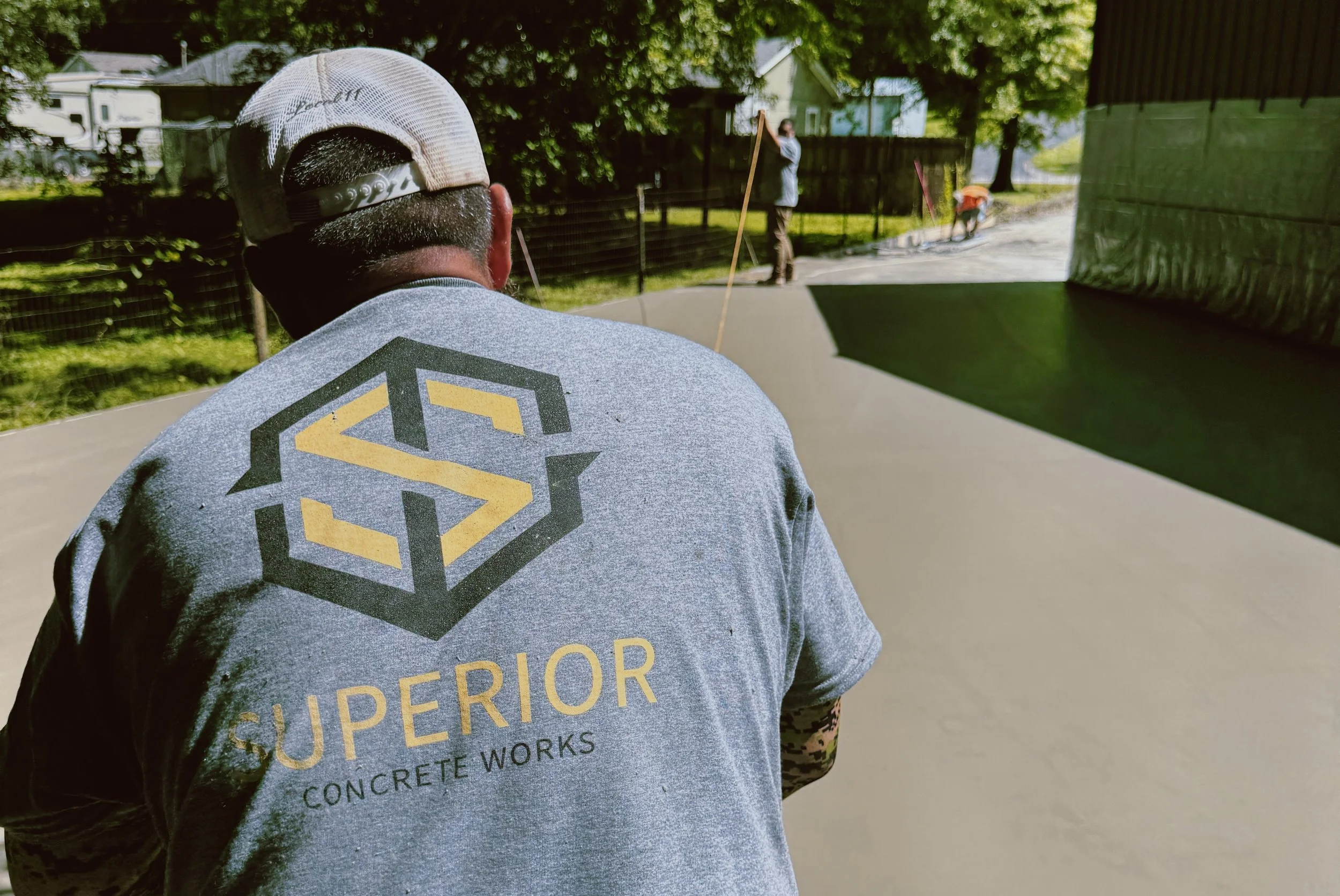 A man wearing a gray sweatshirt with a black and yellow logo on the back, and a backward baseball cap, is kneeling on a freshly poured concrete driveway. In the background, another worker is spreading and smoothing concrete, with a person standing nearby holding a tool. There is greenery and trees, a black fence, and houses in the distance.