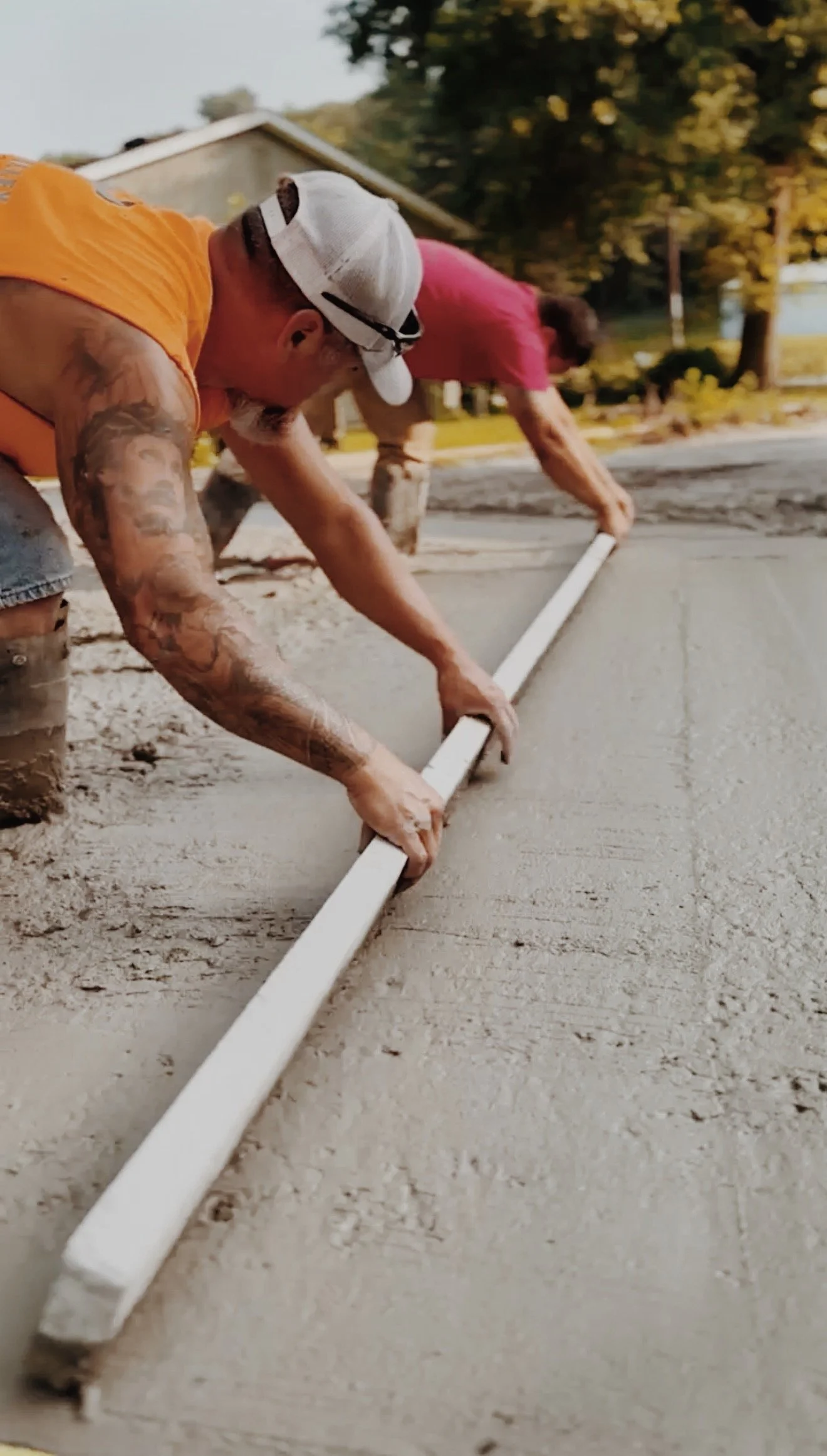 Two construction workers are applying concrete to a sidewalk, using a long white level and wearing work clothes and hats.