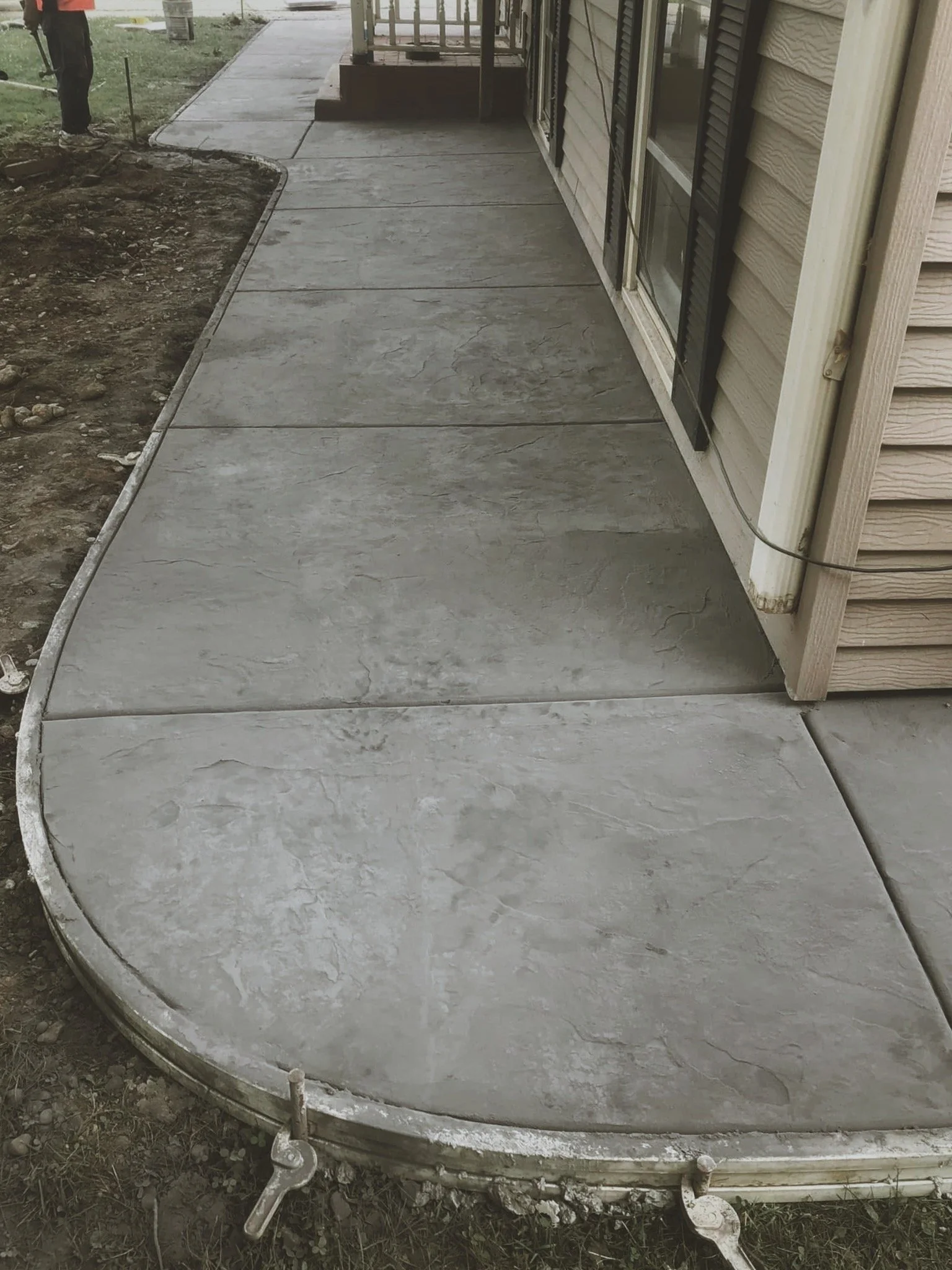 Concrete sidewalk with rounded edges along the side of a house, with some garden bed area on the left side and a person standing in the background.
