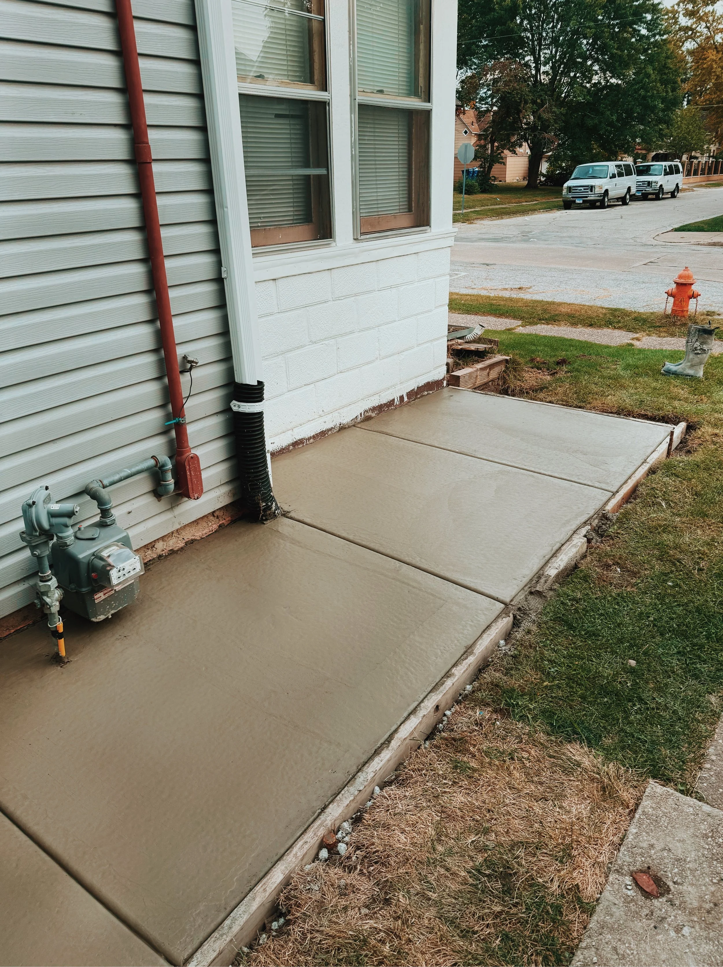 Newly poured concrete sidewalk next to a house with beige siding and a white corner trim, with utility meters and pipes at the base, and a grassy yard with a red fire hydrant and a worn boot nearby, in a neighborhood with trees and parked white vans 