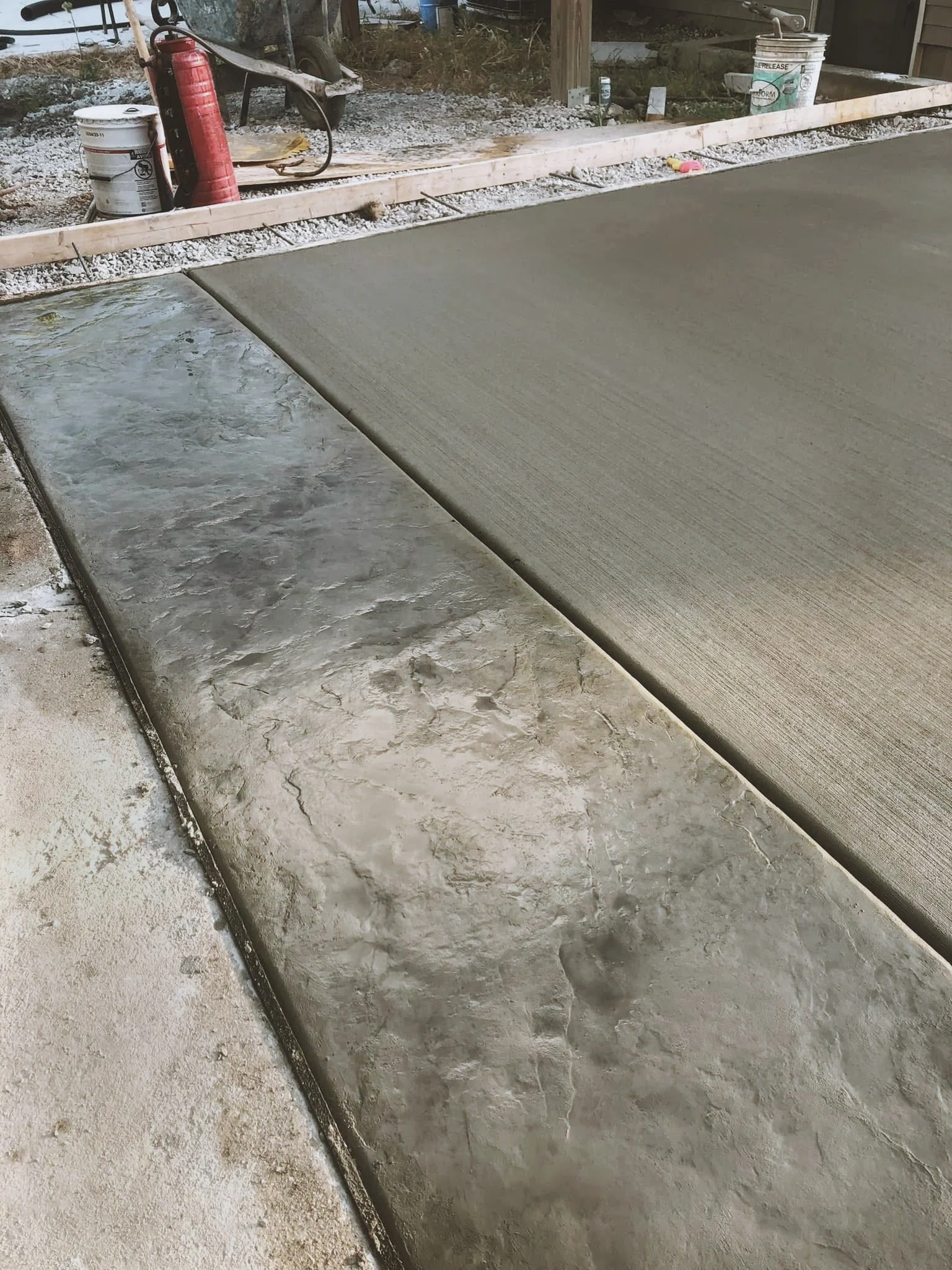 Newly poured concrete sidewalk with a rough surface next to an older, cracked concrete sidewalk. In the background, there are construction tools, an orange fire extinguisher, a wheelbarrow, and buckets.