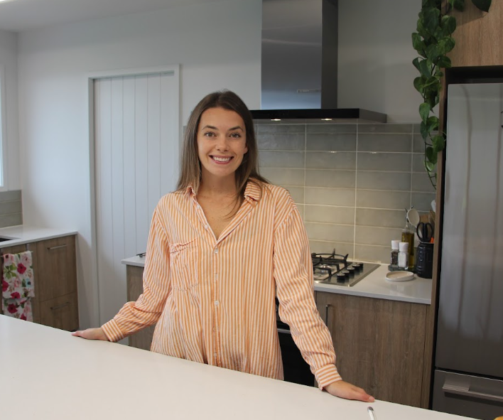 A woman with long hair, smiling, wearing a striped orange and white shirt, standing in a modern kitchen.