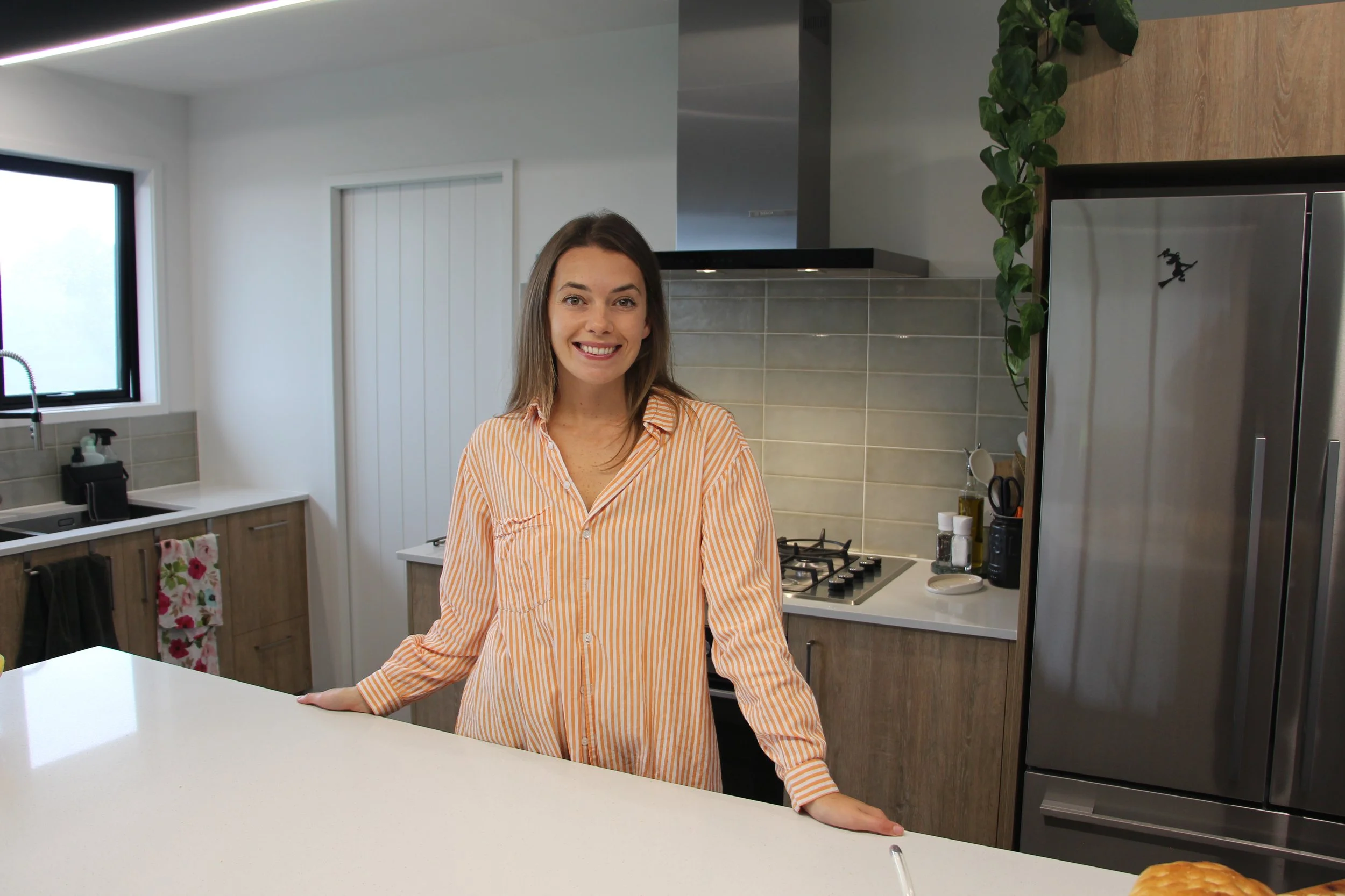 A woman with long brown hair wearing a striped orange and white shirt, smiling and leaning on a kitchen counter in a modern kitchen with a window, stove, and refrigerator.