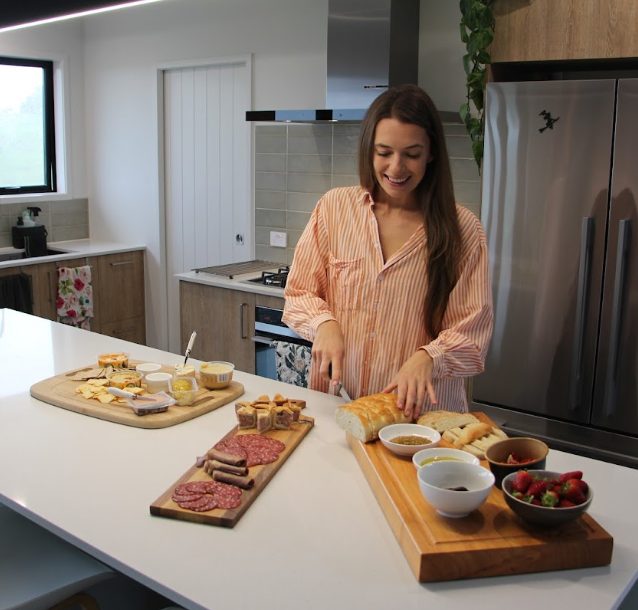 A woman preparing breakfast in a modern kitchen with cheese, meats, bread, strawberries, and spreads on wooden boards.