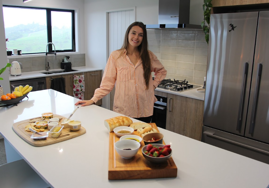 A woman standing in a modern kitchen smiling at the camera, with a table of breakfast foods including bread, strawberries, yogurt, and spreads in front of her.