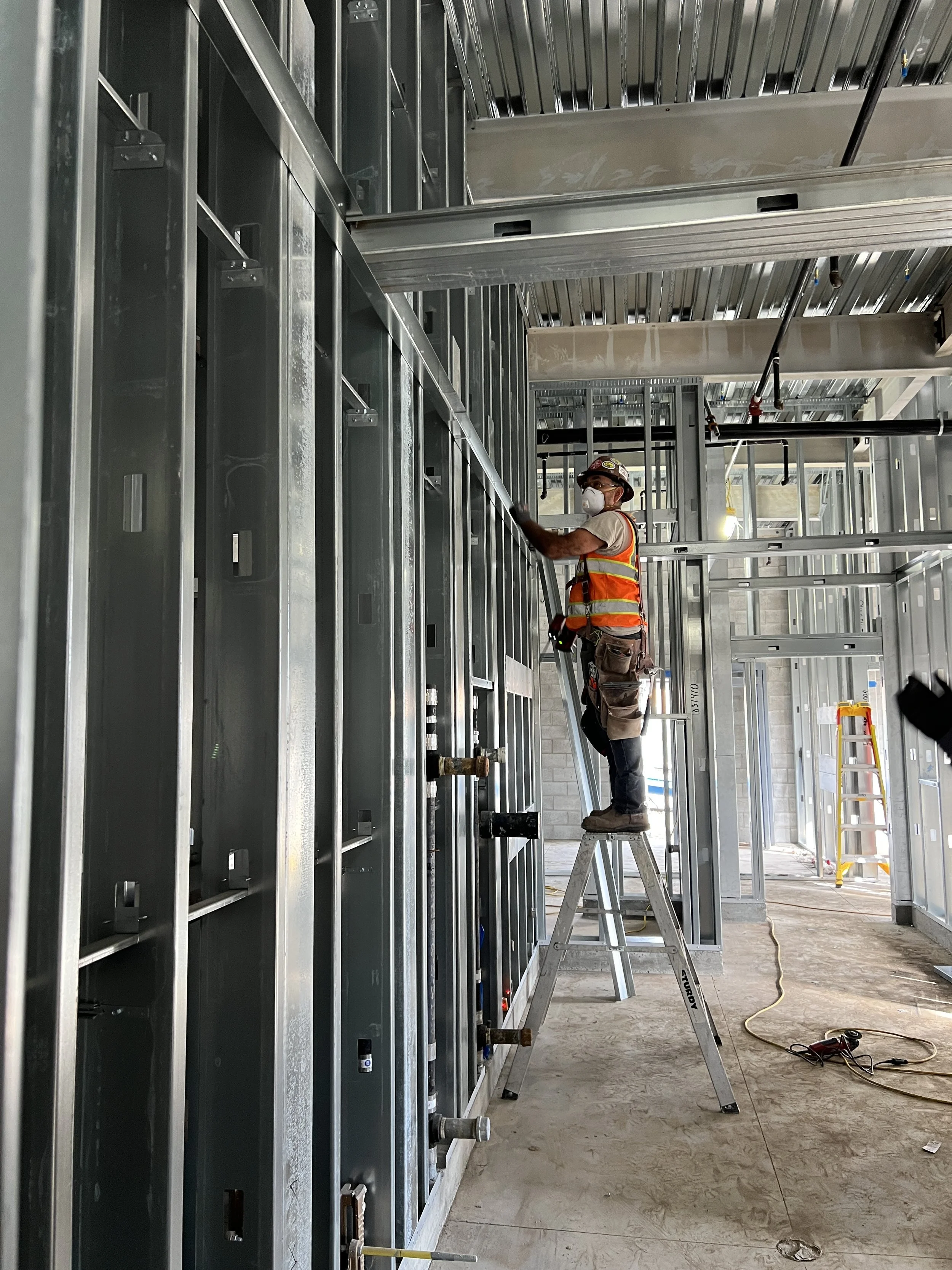 Construction worker wearing a mask, safety vest, and helmet standing on a ladder, working on metal framing inside a building under construction.