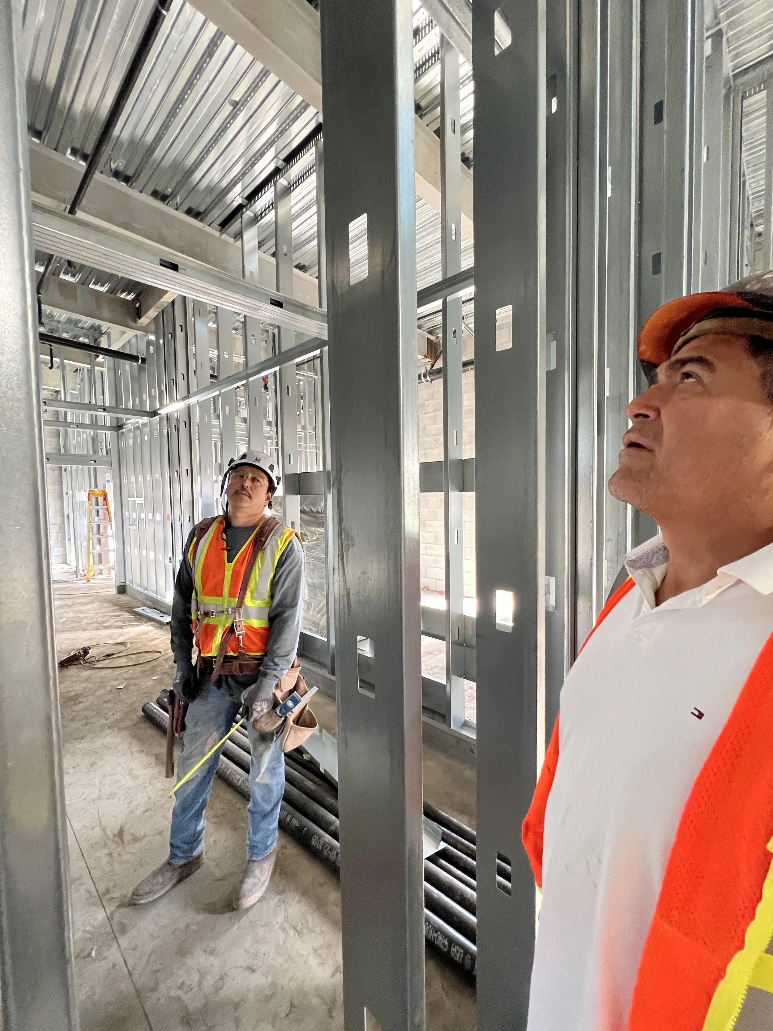Two construction workers inside a building under construction, surrounded by metal framing, with one wearing a white hard hat and the other wearing an orange hard hat, both looking upward.