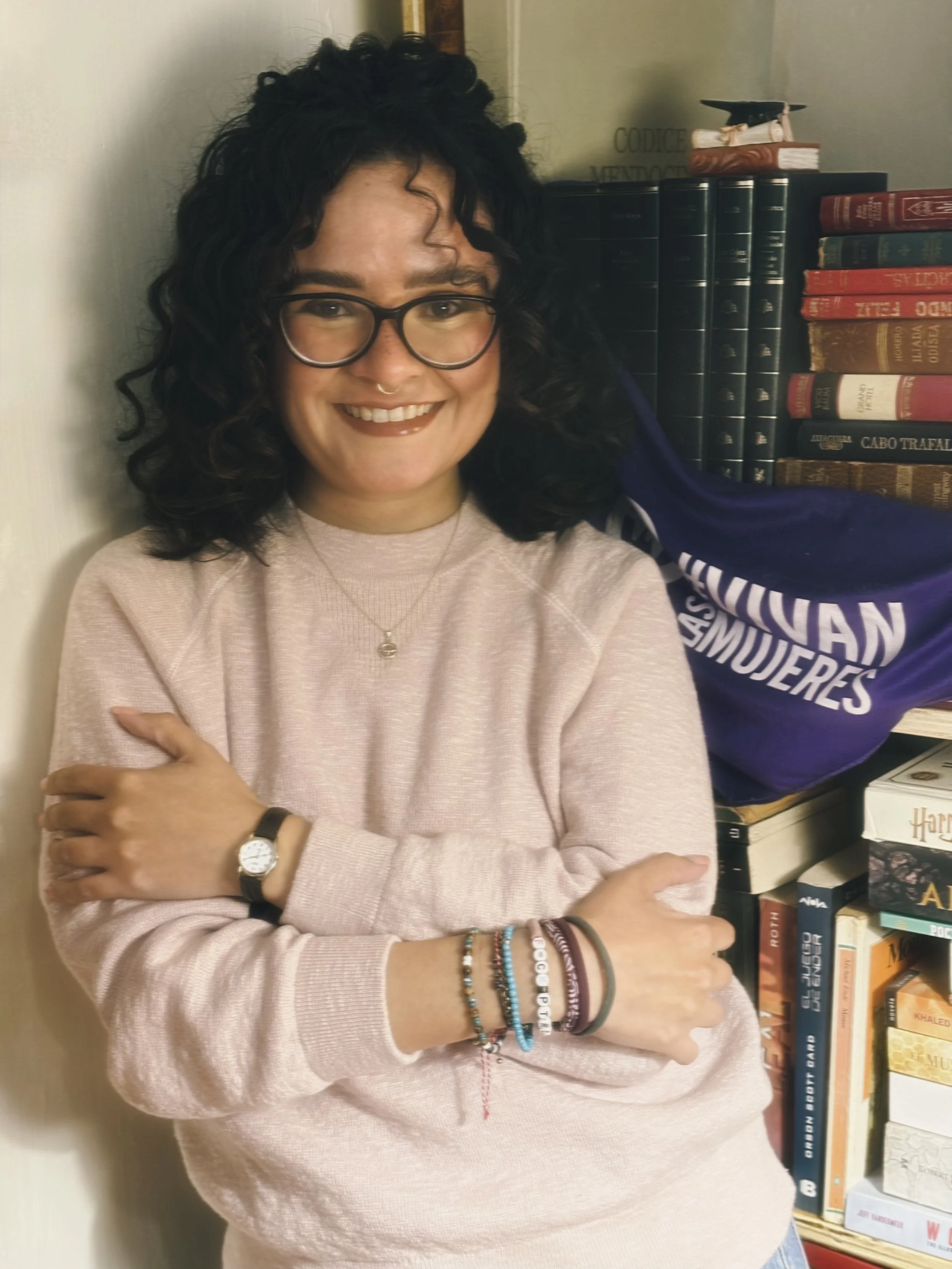 Mujer con gafas, cabello rizado y oscuro, sonriendo. Lleva suéter rosa y varias pulseras, con fondo de libros y bandera de una organización femenina.