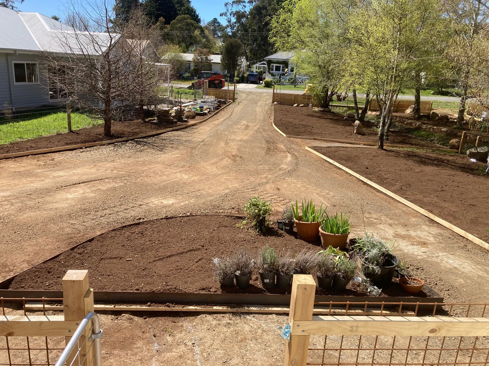 Front yard with newly planted flower beds, potted plants, and a dirt pathway, with trees, houses, and a street in the background.