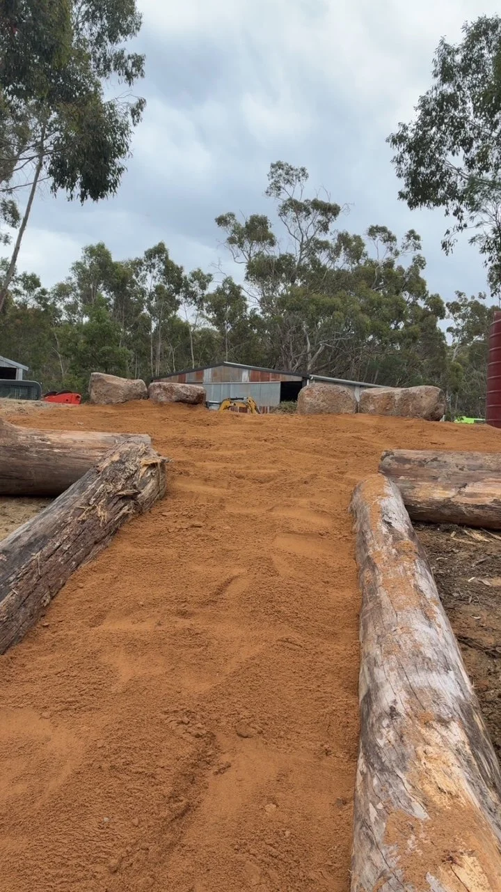 A dirt pathway with logs on either side, leading up to large rocks and a metal building in the background, surrounded by trees under a cloudy sky.