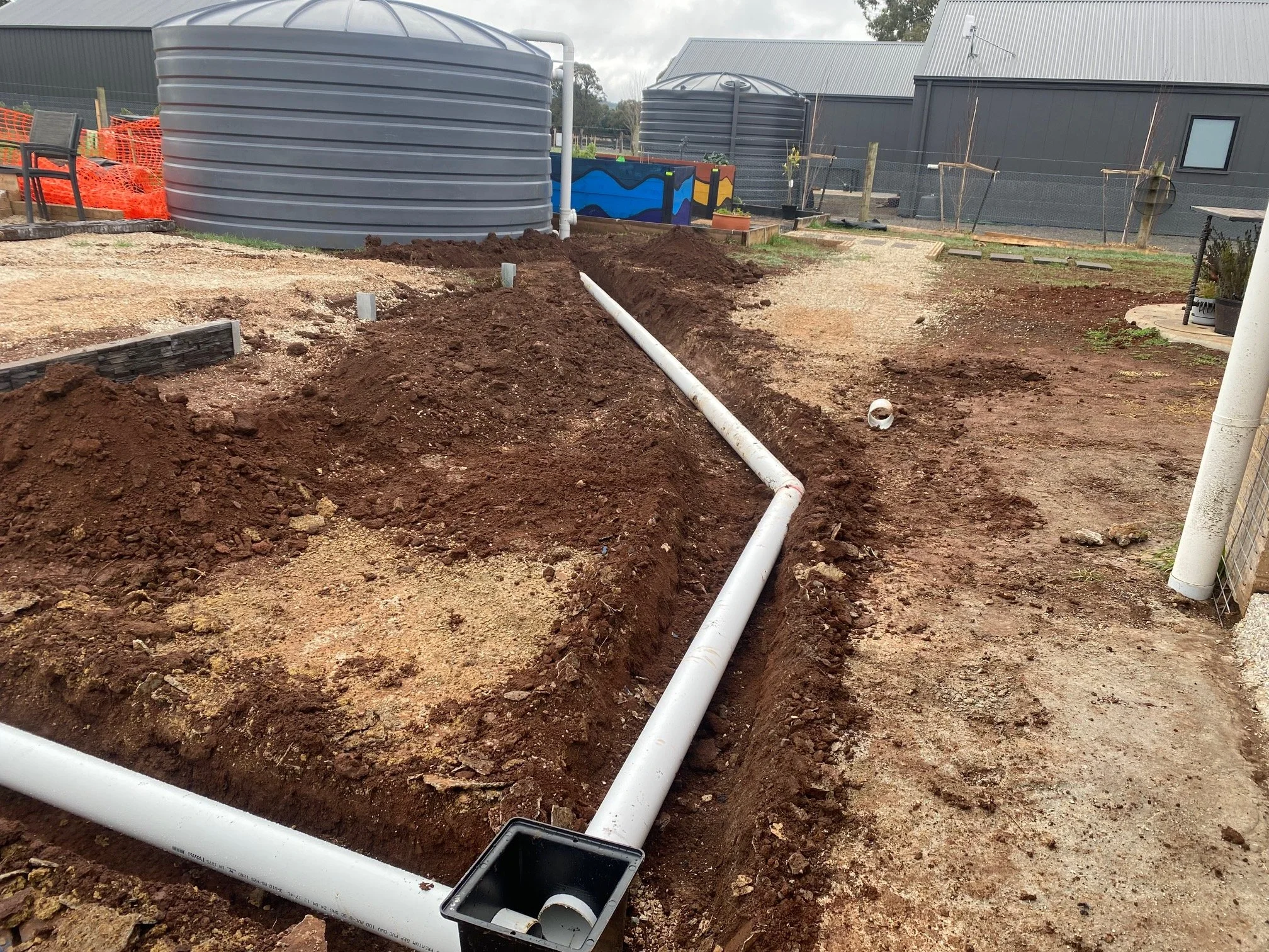 Construction site with underground pipe installation, dirt trench with white pipes, water tanks, and a building in the background.