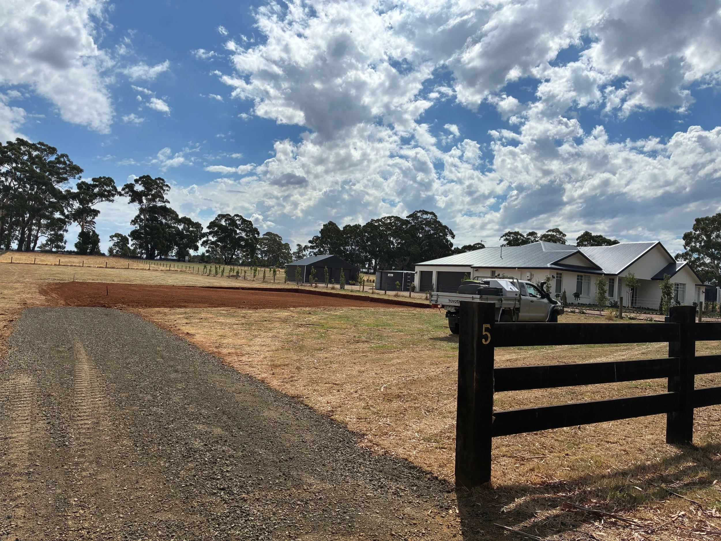A rural property with a house under construction, a dirt and gravel driveway, a pickup truck parked near the house, a black fence, and trees in the background under a partly cloudy sky.