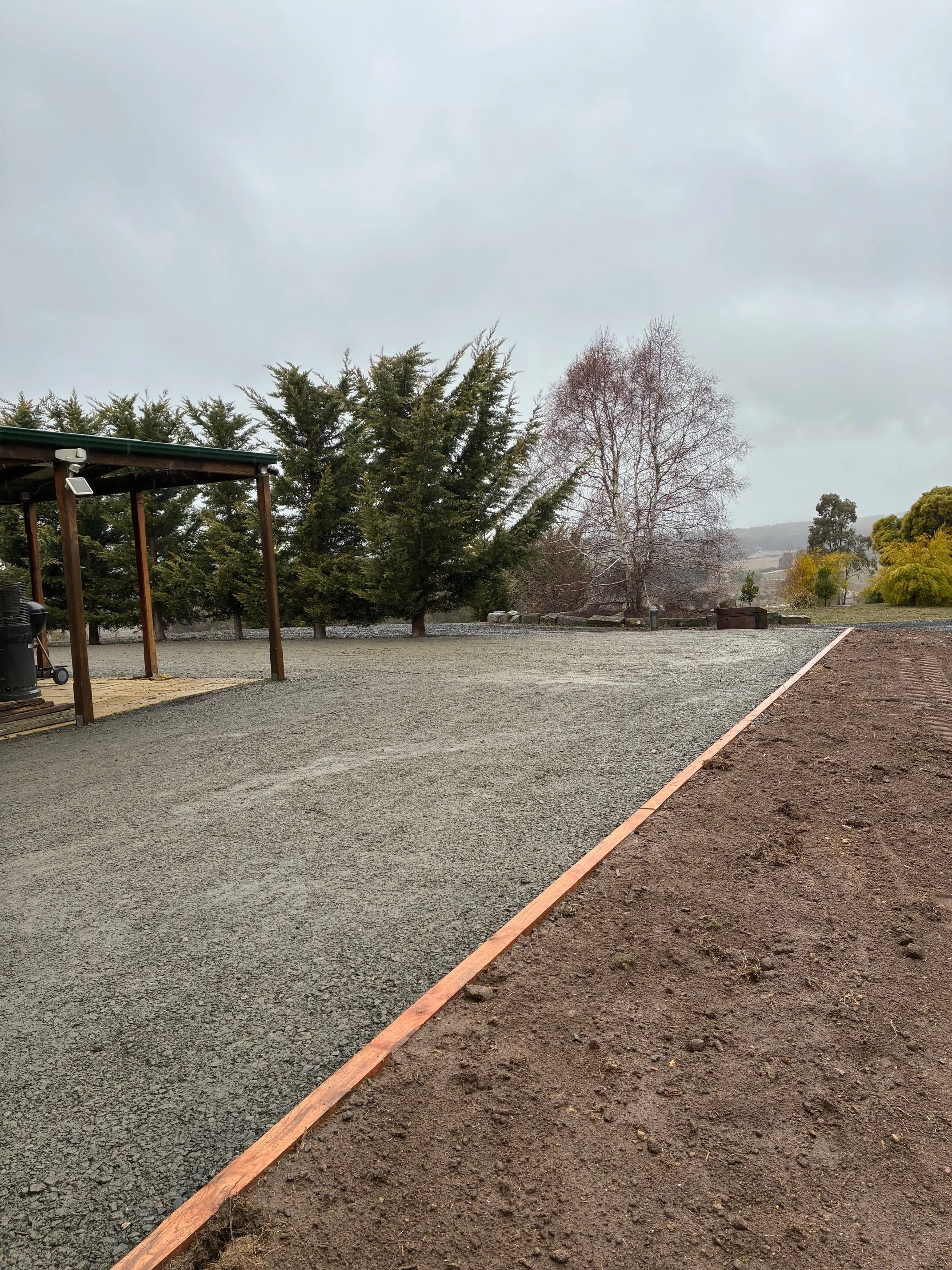An outdoor scene with a gravel pathway bordered by wooden planks, with trees in the background and a cloudy sky overhead.