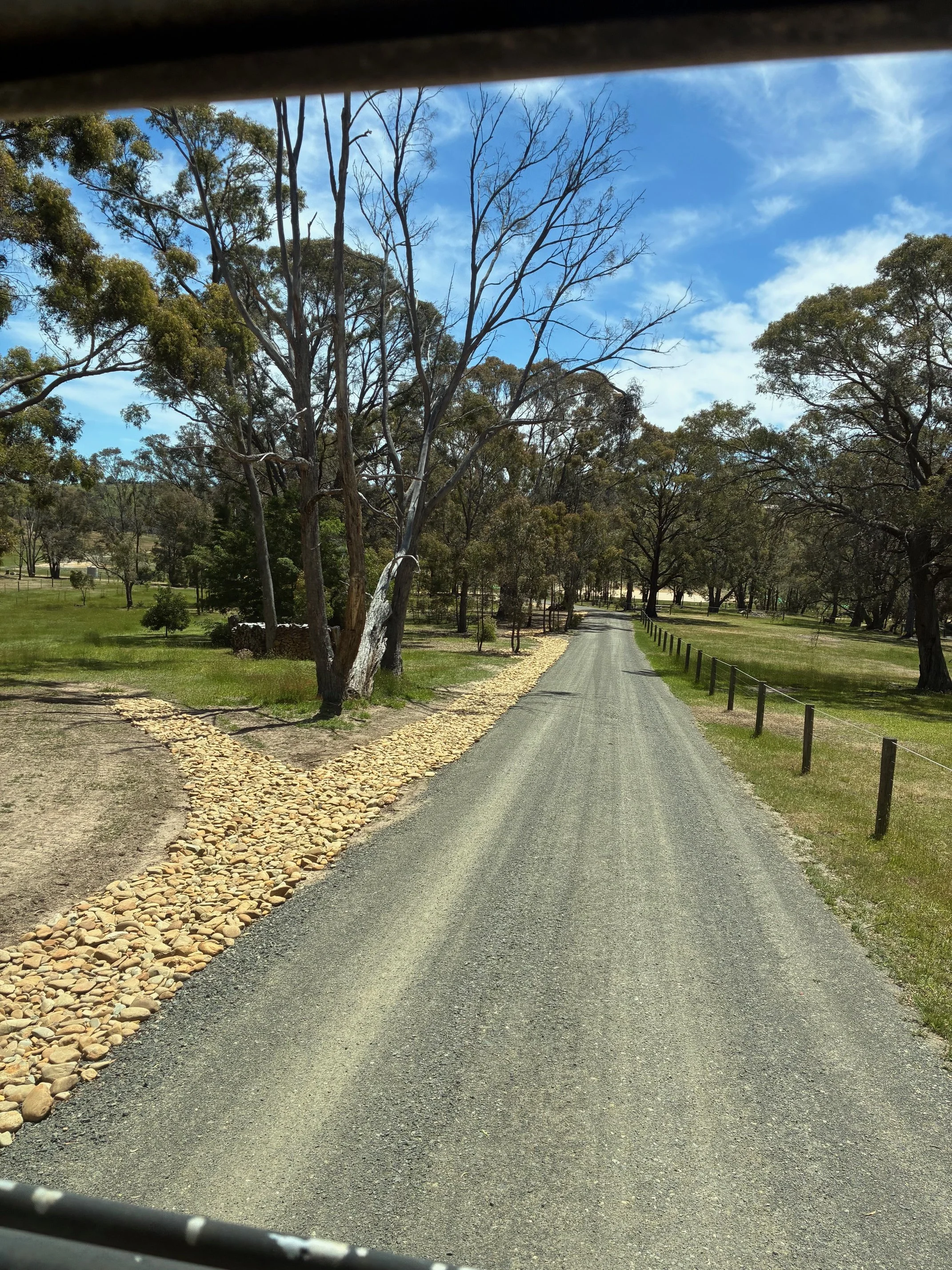 A rural gravel road flanked by trees and grassy areas, with a wood fence on the right side and a stone-lined path on the left, under a partly cloudy blue sky.