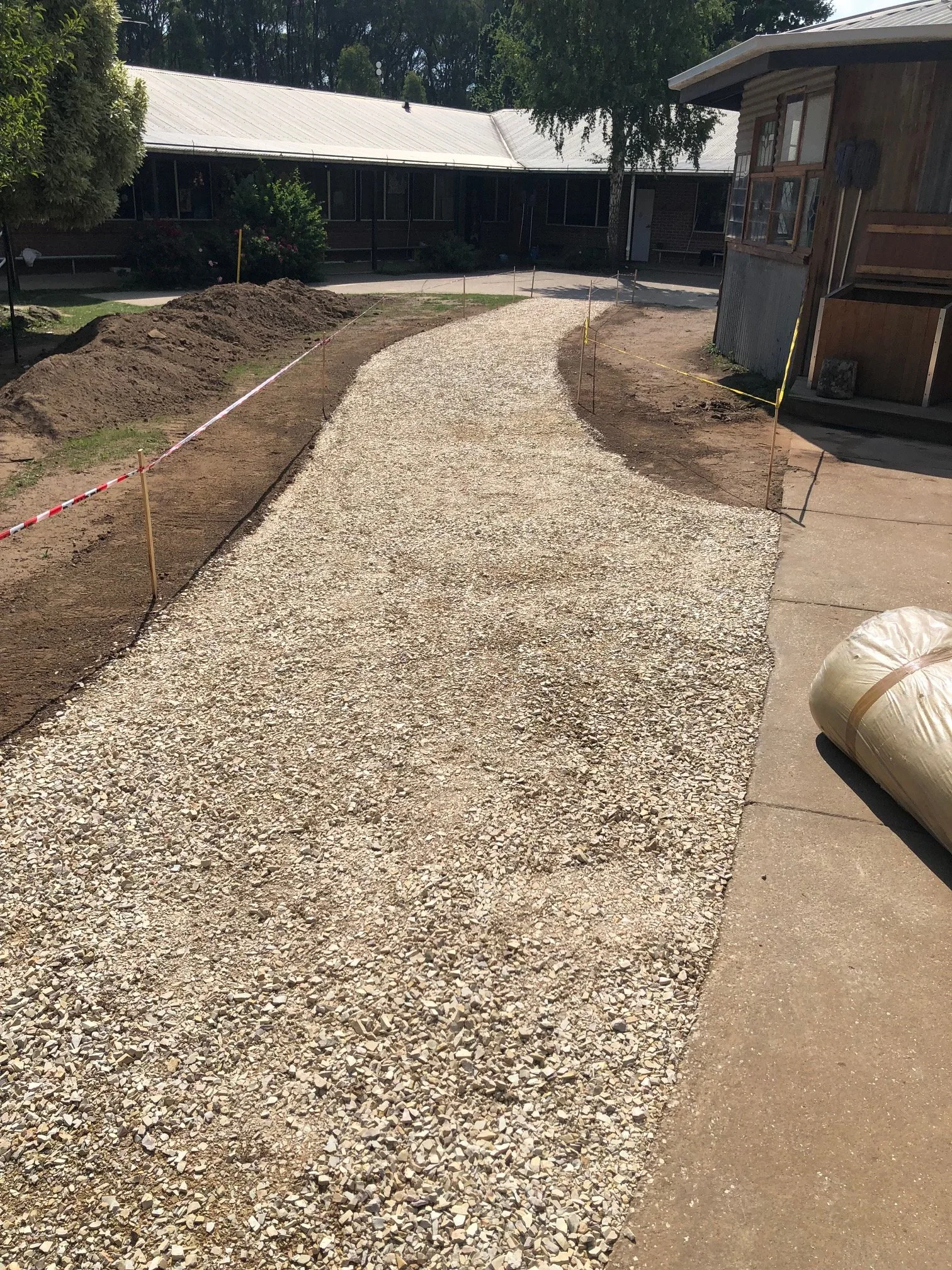 A gravel pathway under construction, bordered by wooden stakes and caution tape, with a building on the right and a house with a metal roof in the background.