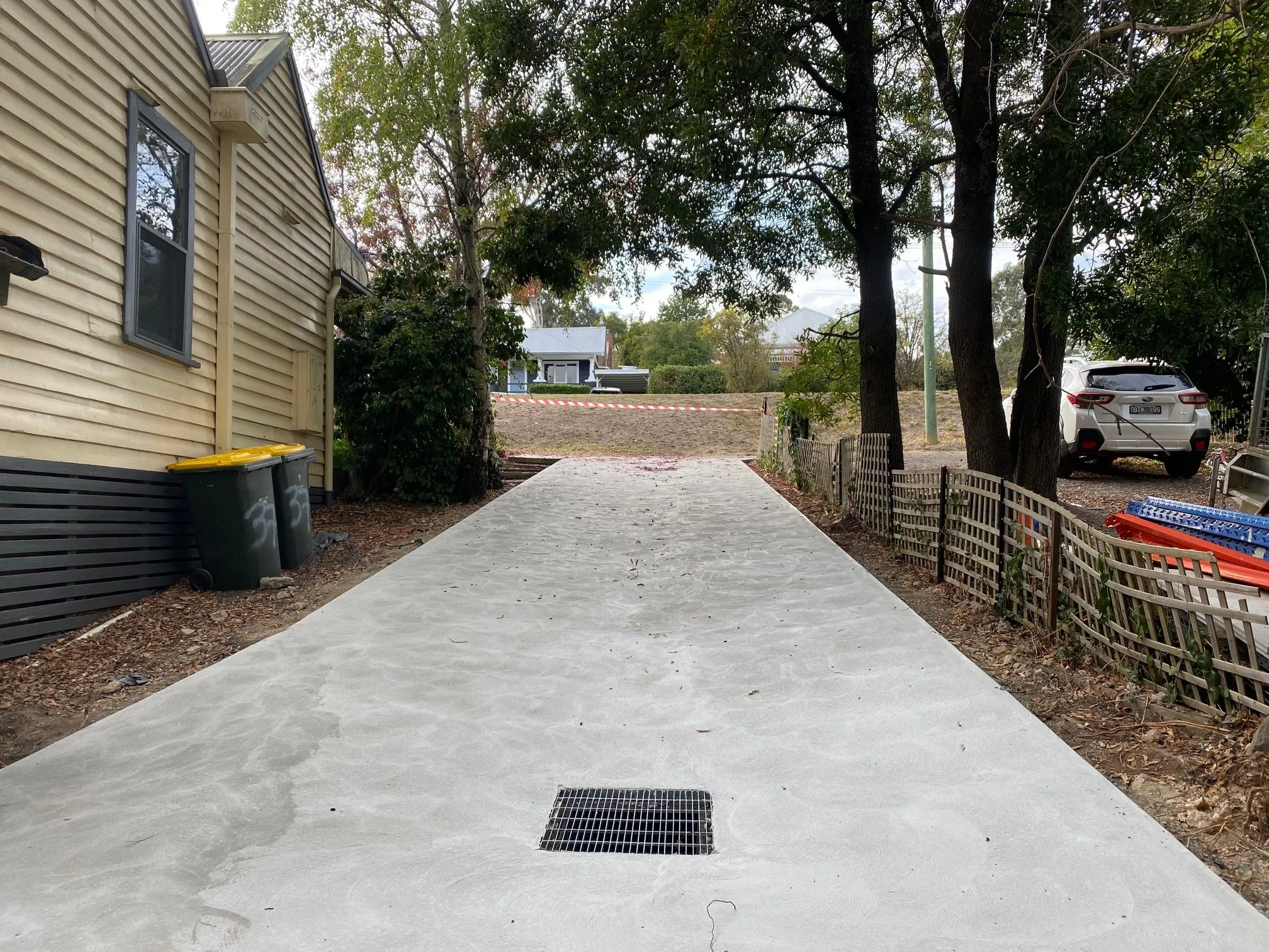 New concrete driveway with drainage grate, side of house with yellow siding, trash bins, parked car, trees, and construction barrier in the background.