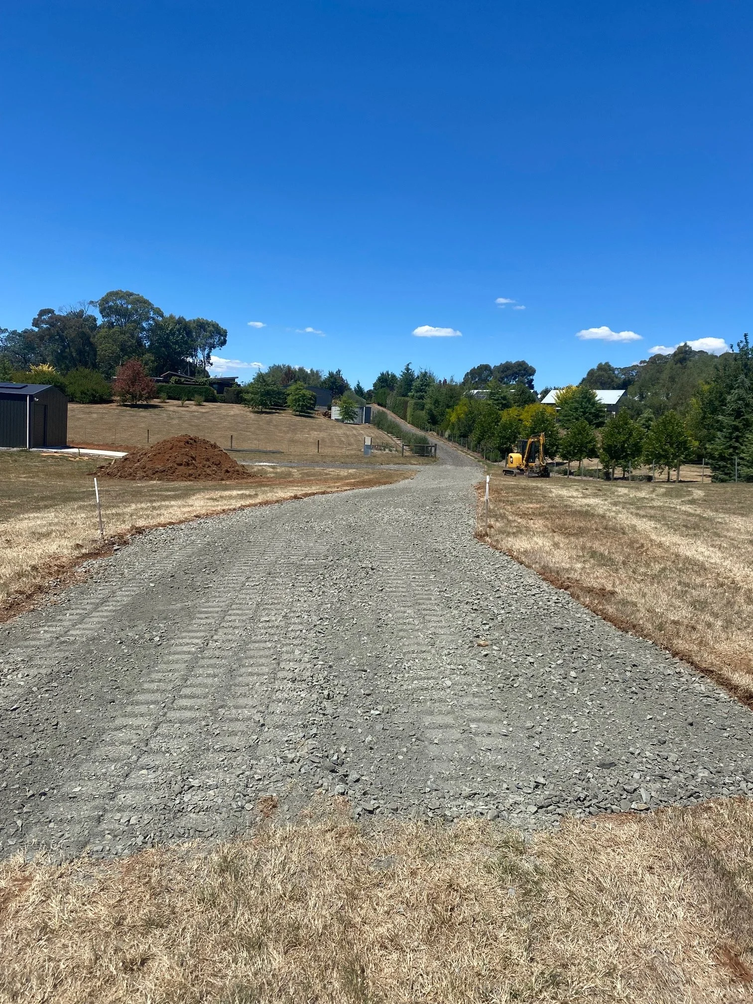 A gravel driveway on a construction site with a yellow excavator on the right side, surrounded by dry grass and trees, under a clear blue sky with a few clouds.
