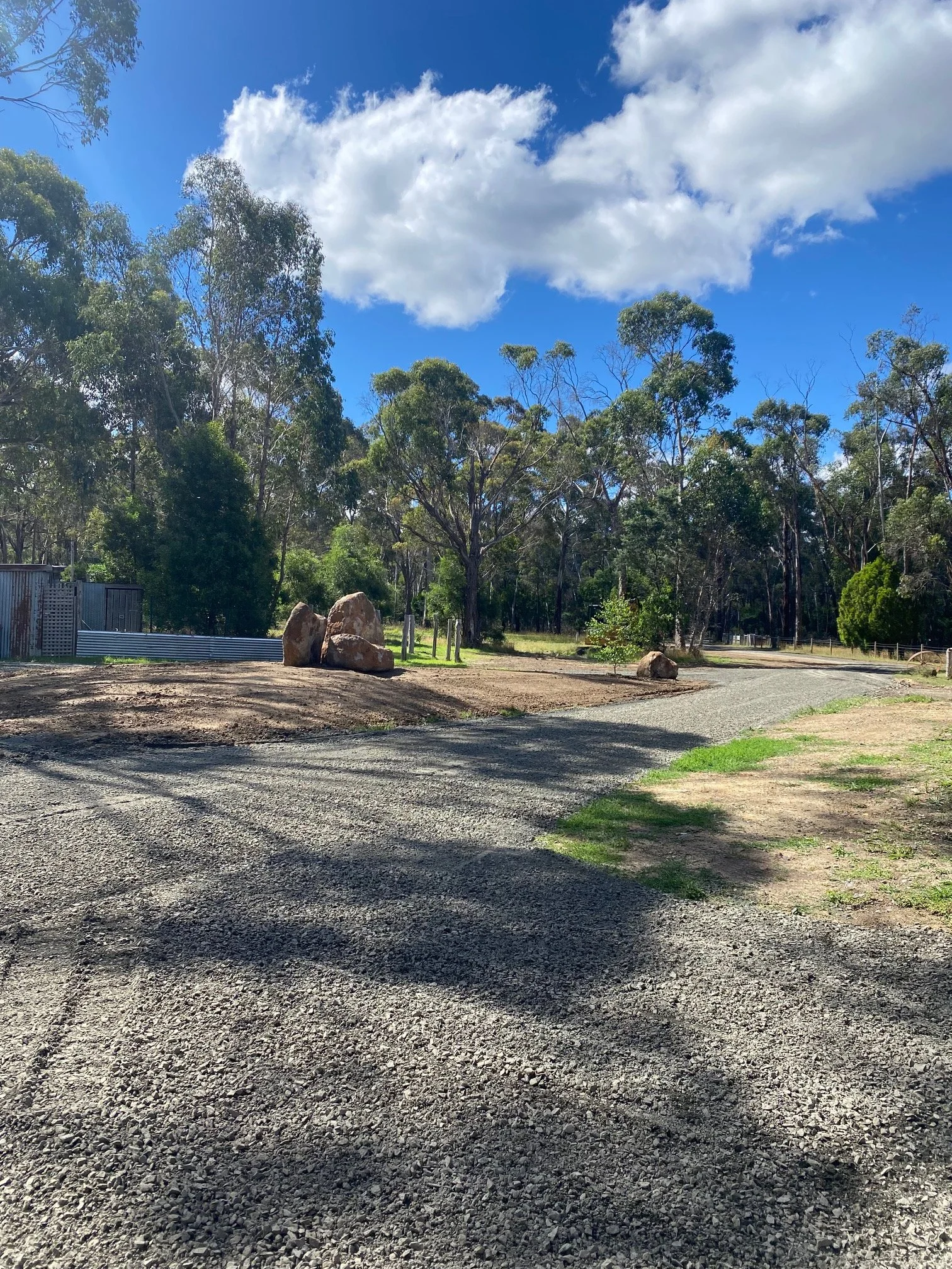 A gravel road curves through a wooded area with tall trees, a blue sky with clouds overhead, and a few large rocks on the side of the road.